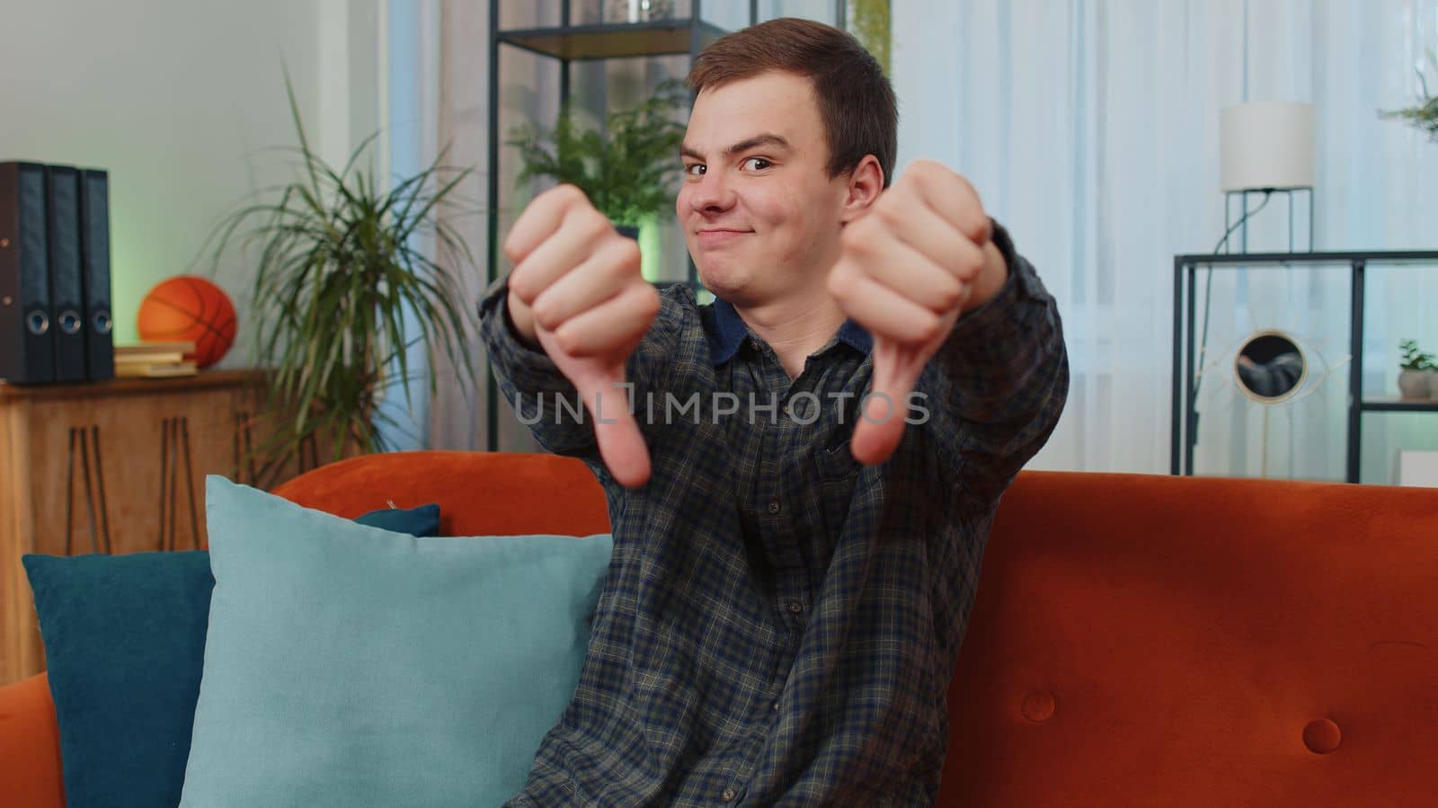 Dislike. Upset teen man showing thumbs down sign gesture, expressing discontent, disapproval, dissatisfied bad loser work at modern home apartment indoors. Displeased young guy in living room on sofa