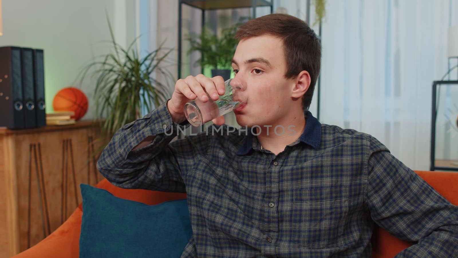 Portrait of thirsty teenager man sitting indoors holding glass of natural aqua make sips drinking still water preventing dehydration. Guy with good life habits, healthy slimming, weight loss concept