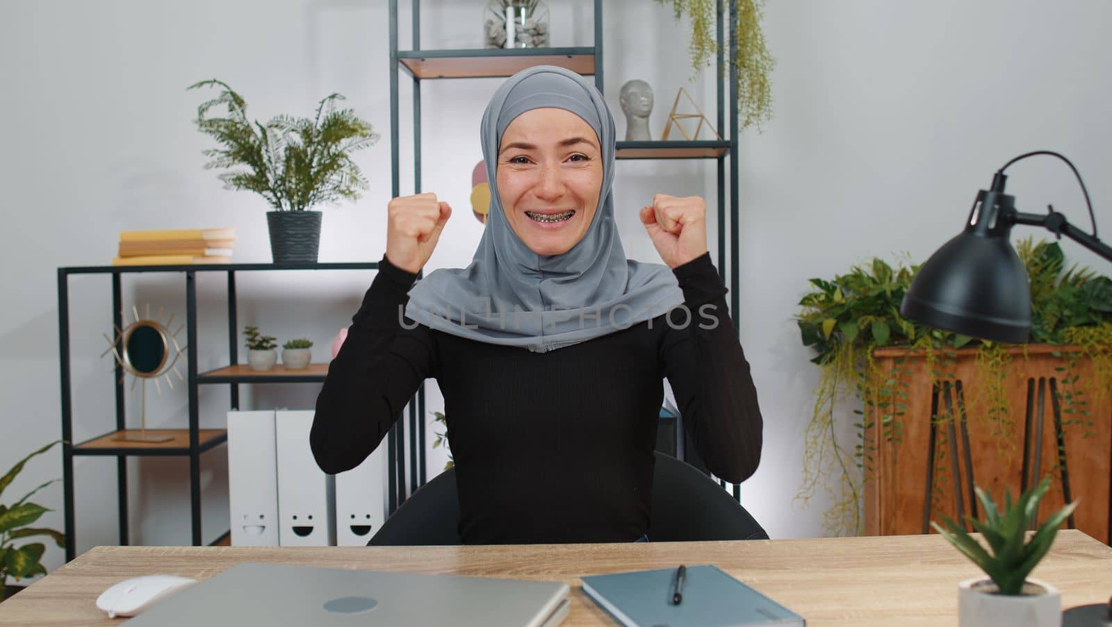 Happy young muslim business woman wearing hijab headscarf shouting, celebrating success victory, winning, goal achievement, good news, lottery jackpot luck, surprise. Girl at home office workspace