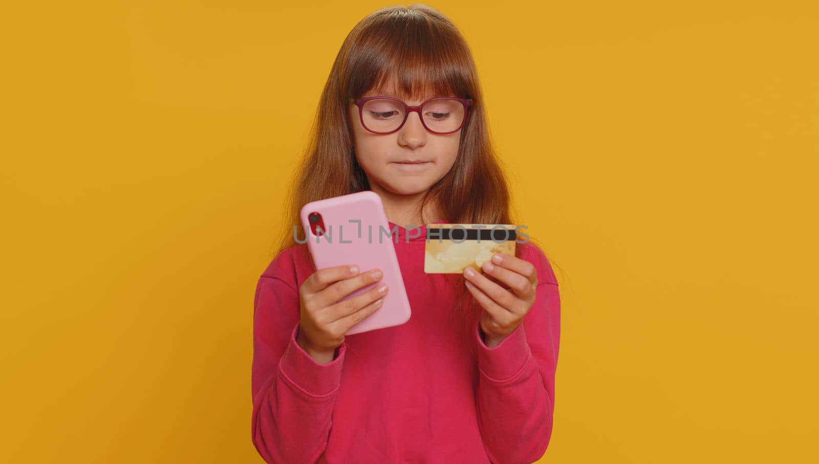 Young preteen child girl kid using credit bank card and smartphone while transferring money, purchases online shopping, order food delivery. Little toddler children alone on studio yellow background