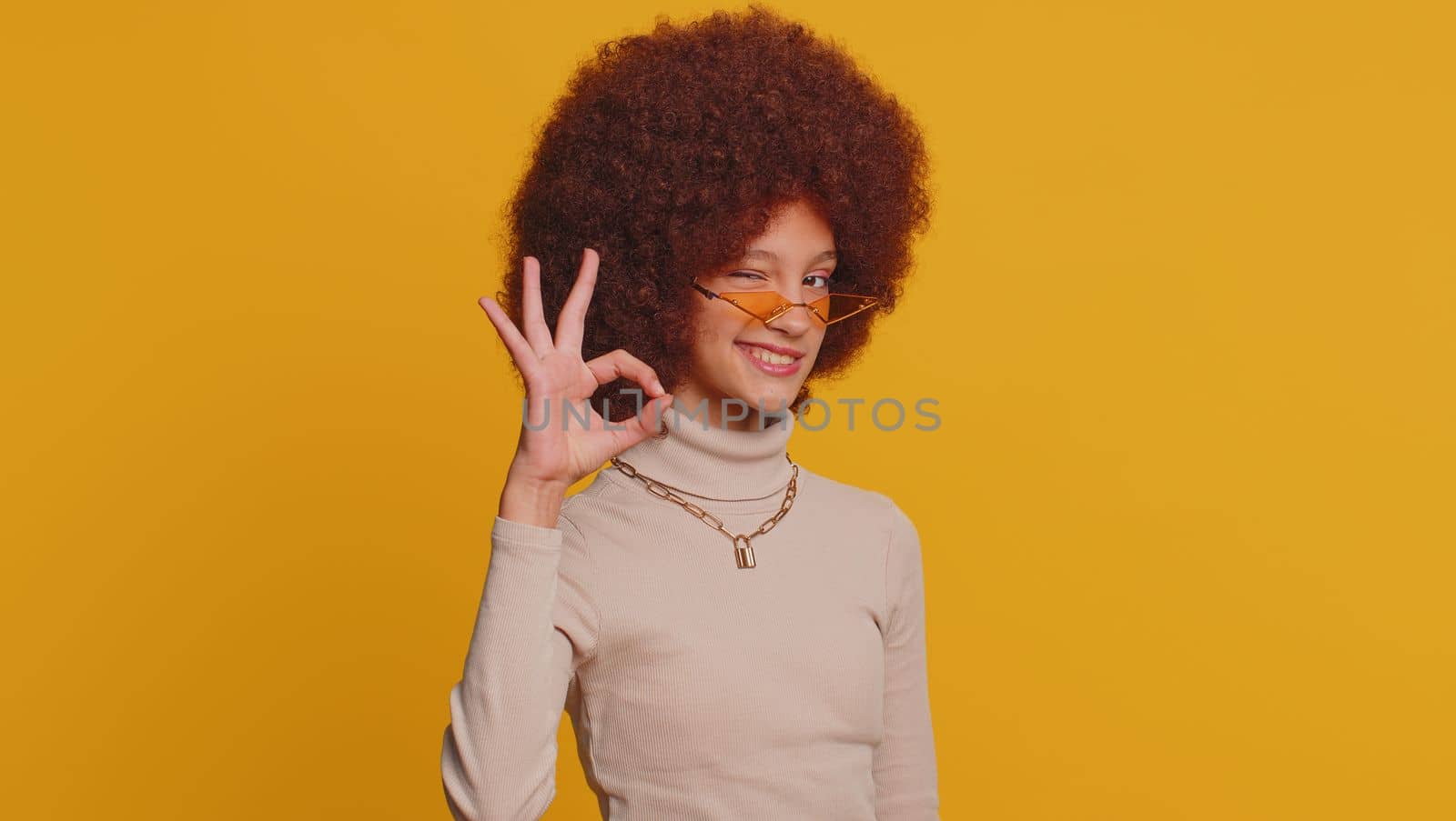 Ok. Young funny school girl in sunglasses looking approvingly at camera showing okey gesture, like sign positive, approve something good. Teenager female child kid isolated on studio yellow background