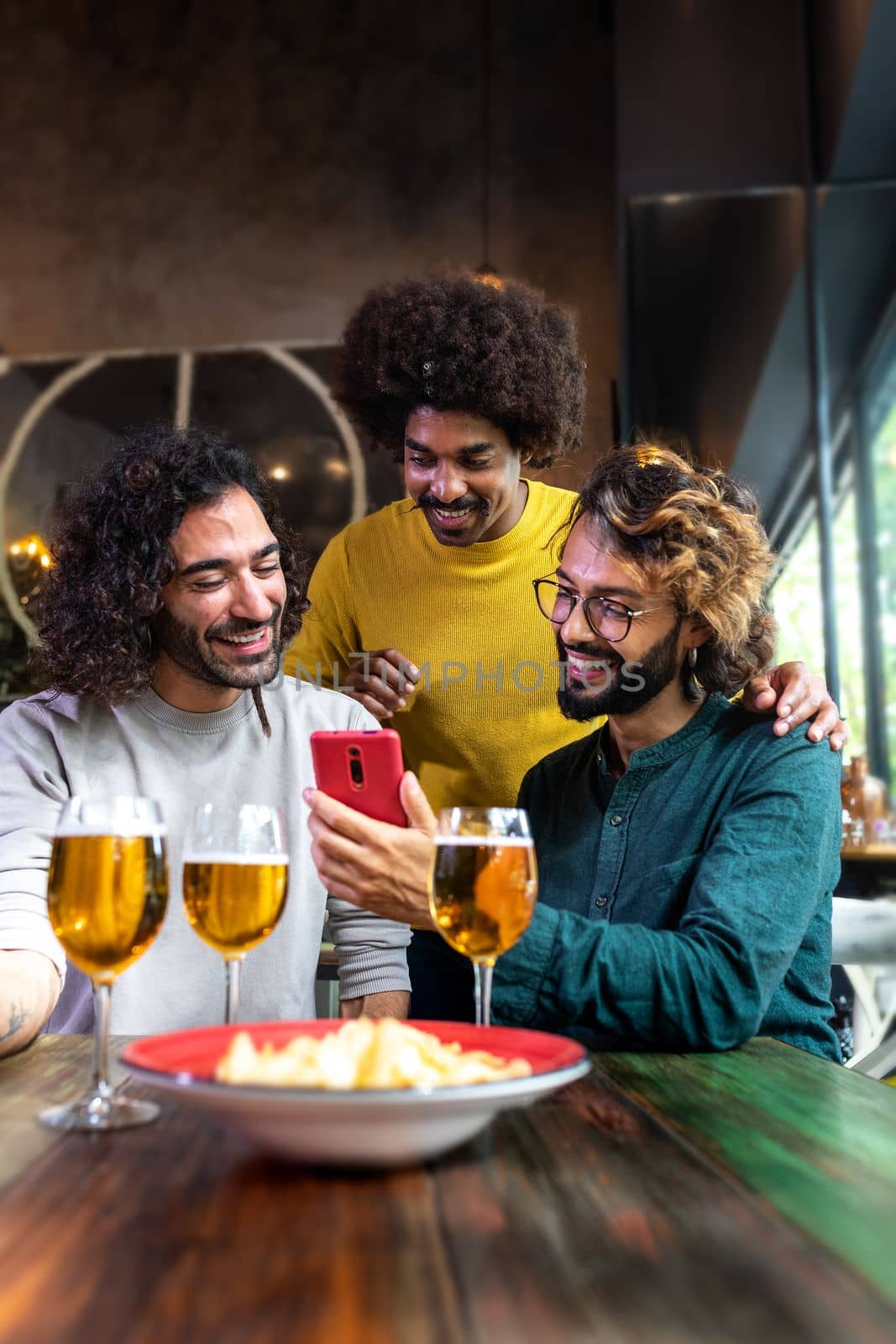 Young man showing to male friends mobile phone while having drinks together in pub. Vertical image.Technology and leisure activity.