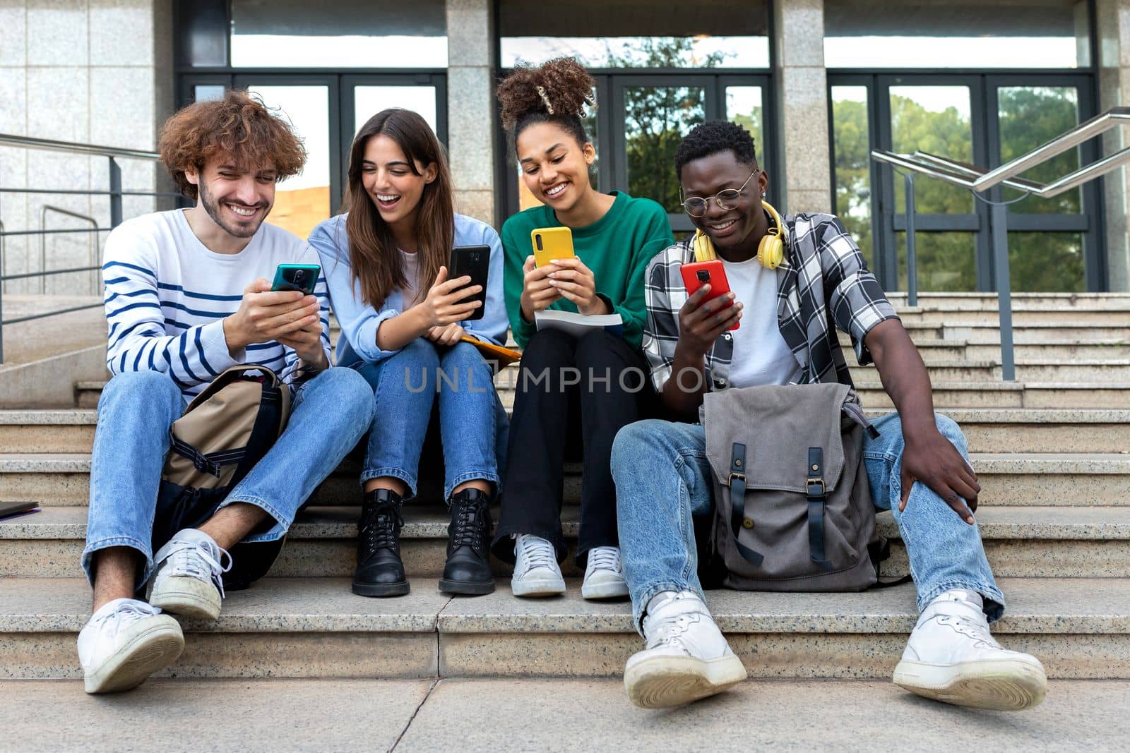 Multiracial college student friends look mobile phone laughing together. University students using smartphone outdoors. by Hoverstock