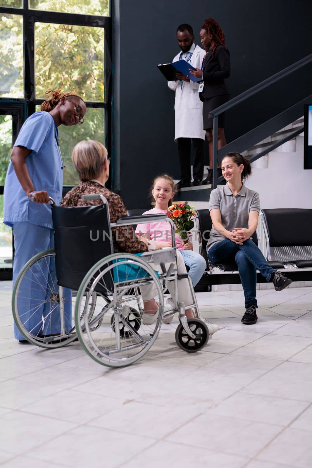 Medical assistant bringing old patient to family after finishing appointment while explaining health care treatment in hospital waiting area. Granddaughter holding bouquet of flowers for grandmother