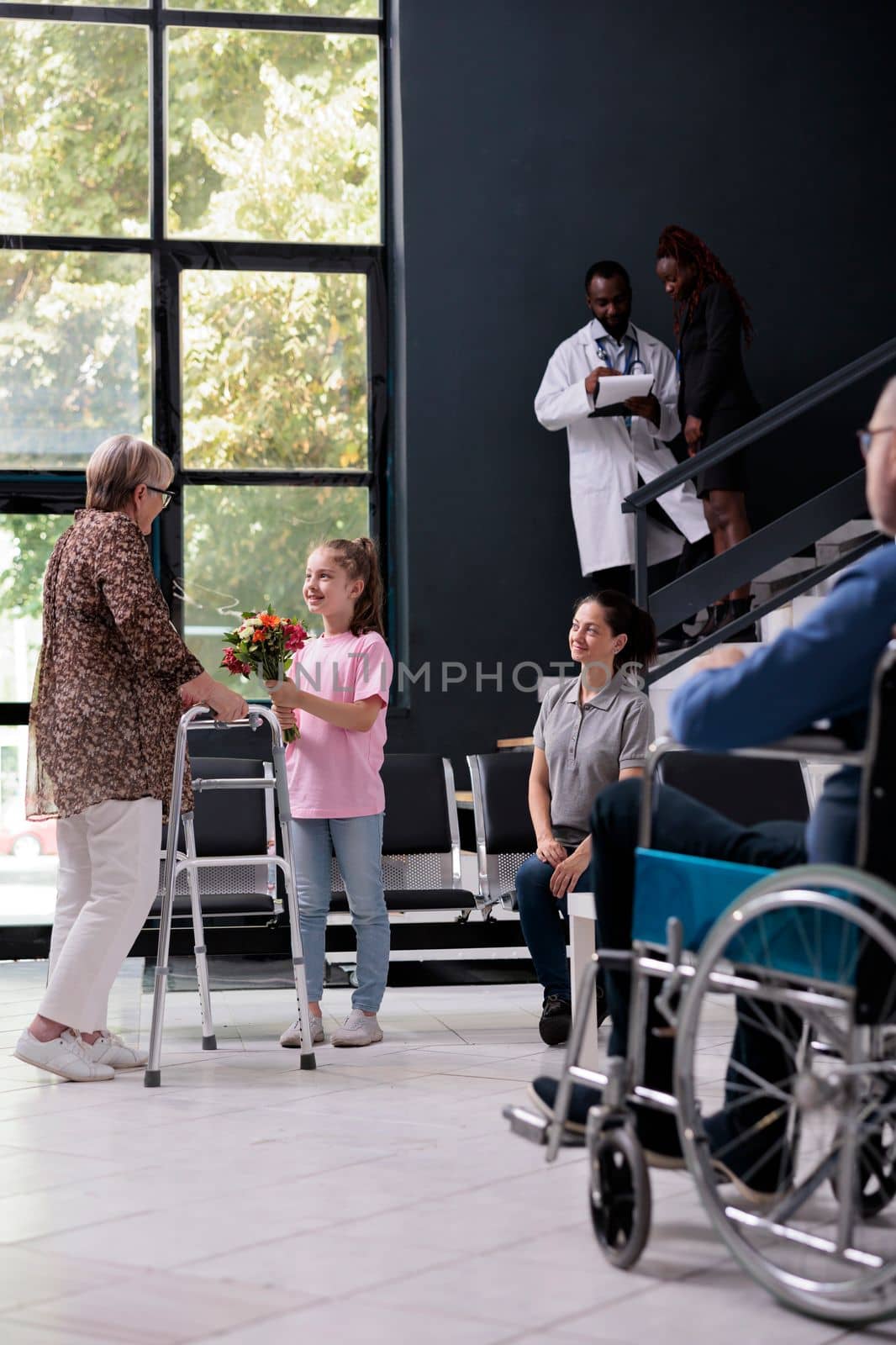 Elderly patient with walking frame standing in hospital reception during checkup visit consultation. Granddaughter welcome her grandmother with bouquet of flowers. Medicine service and concept