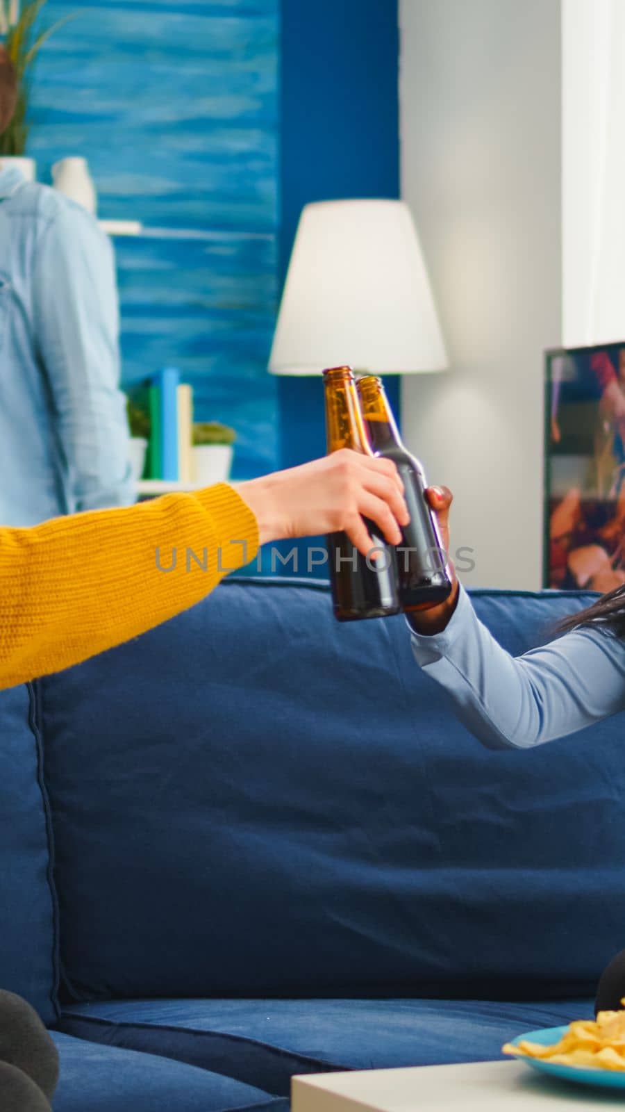 Mixed race black woman taking off protection masks clinking bottles of beer, drinking and eating snacks spending free time in living room respecting social distance. Diverse people enjoying party in outbreak