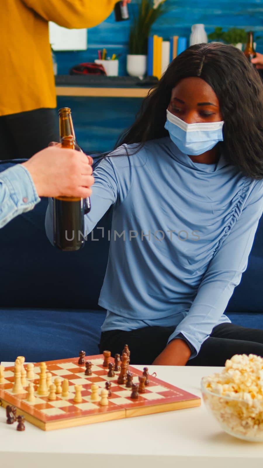 Man holding beer bottle playing chess with african woman in living room keeping social distancing due to covid19 pandemic wearing face mask to prevent infection. Diverse people enjoying board games