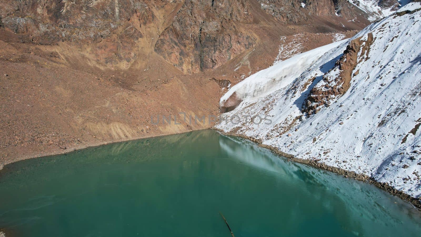 A mountain lake with emerald water reflects a glacier like a mirror. You can see the peaks of the mountains. The lake is partially frozen. There are large stones and snow in places. Moraine Lake