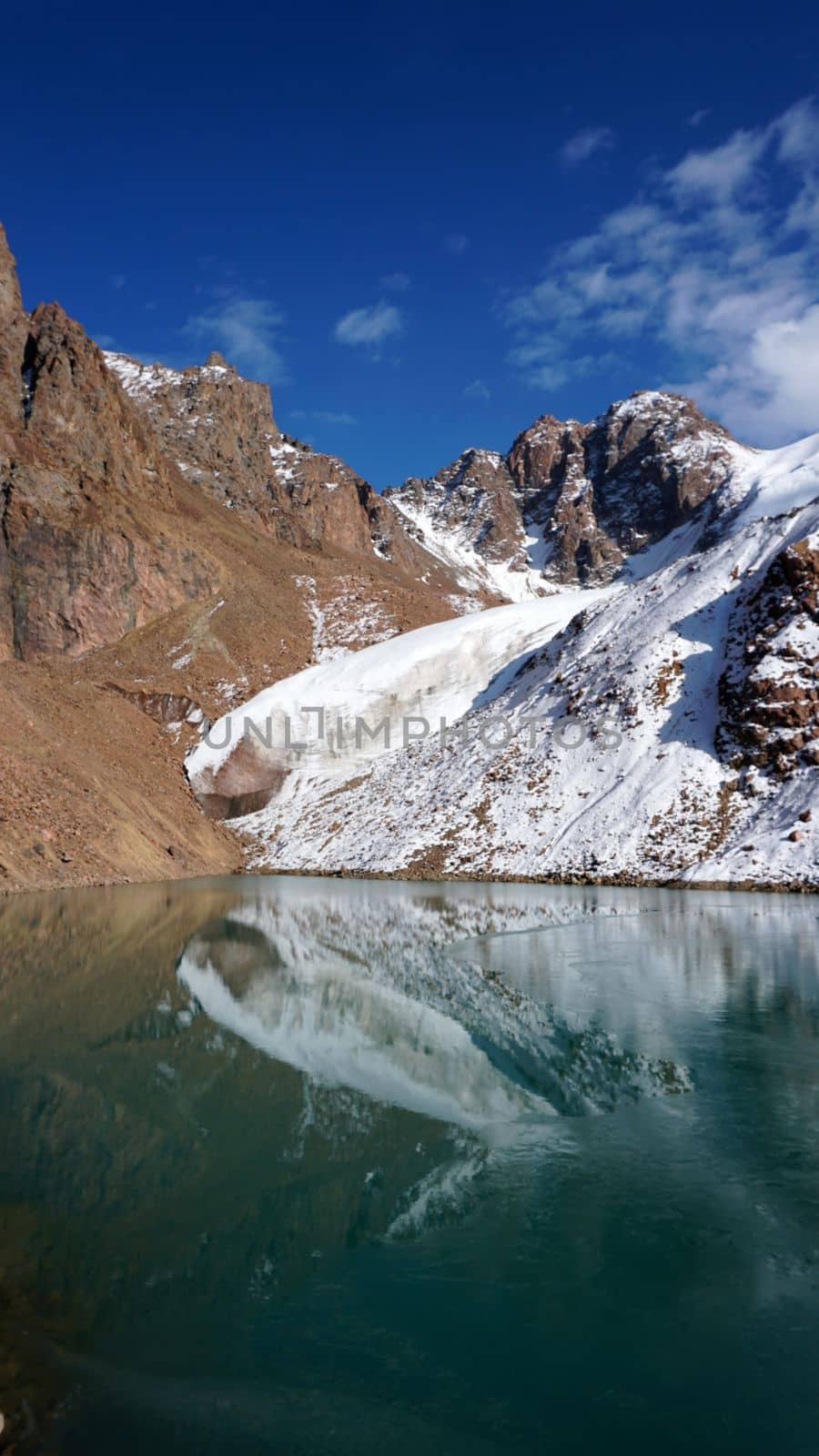 A mountain lake with emerald water reflects a glacier like a mirror. You can see the peaks of the mountains. The lake is partially frozen. There are large stones and snow in places. Moraine Lake