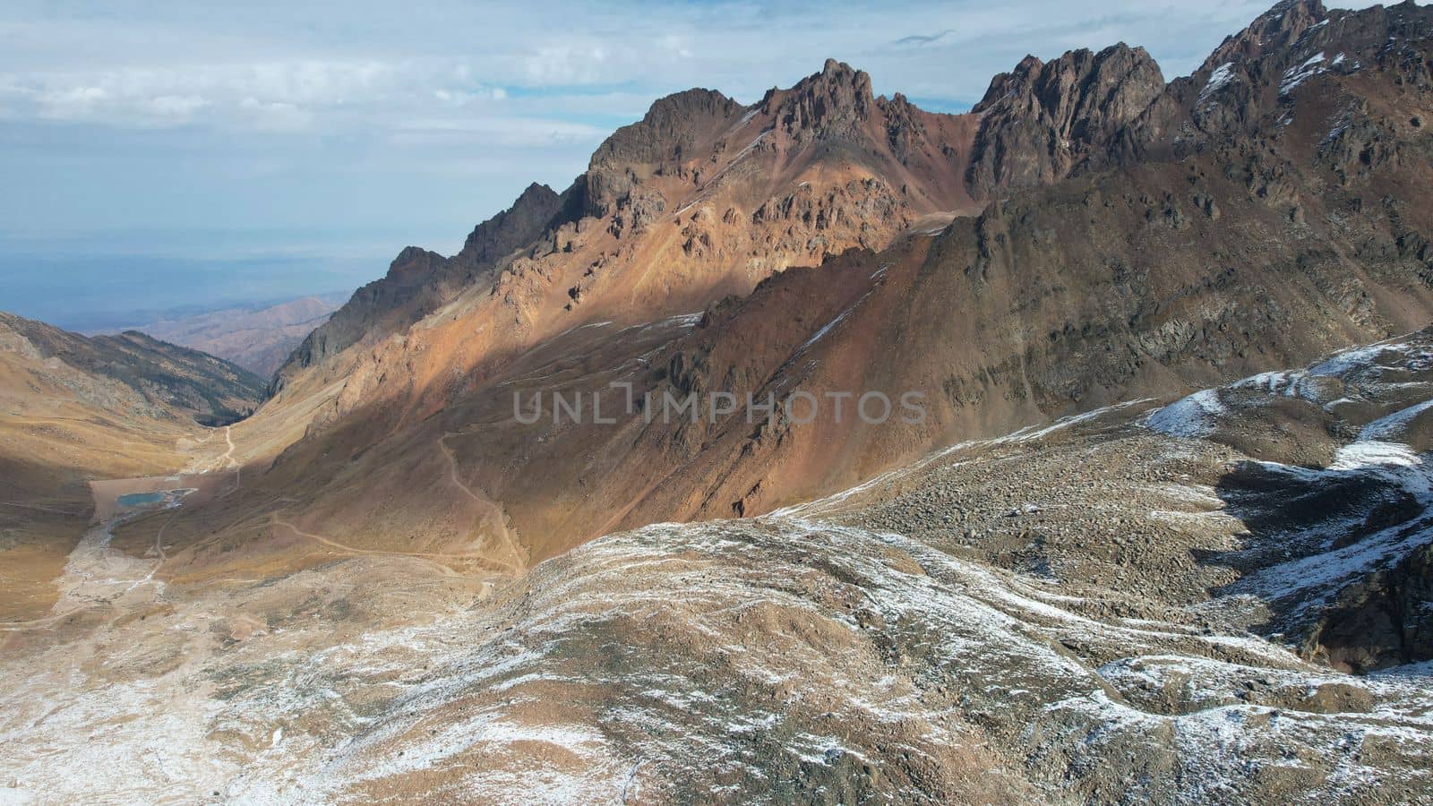 Top view of the high rocky mountains with trails. In places there is snow and yellow-orange plants grow. Shadows from clouds float on the rocks. A place to climb to the top. The mountains of Almaty