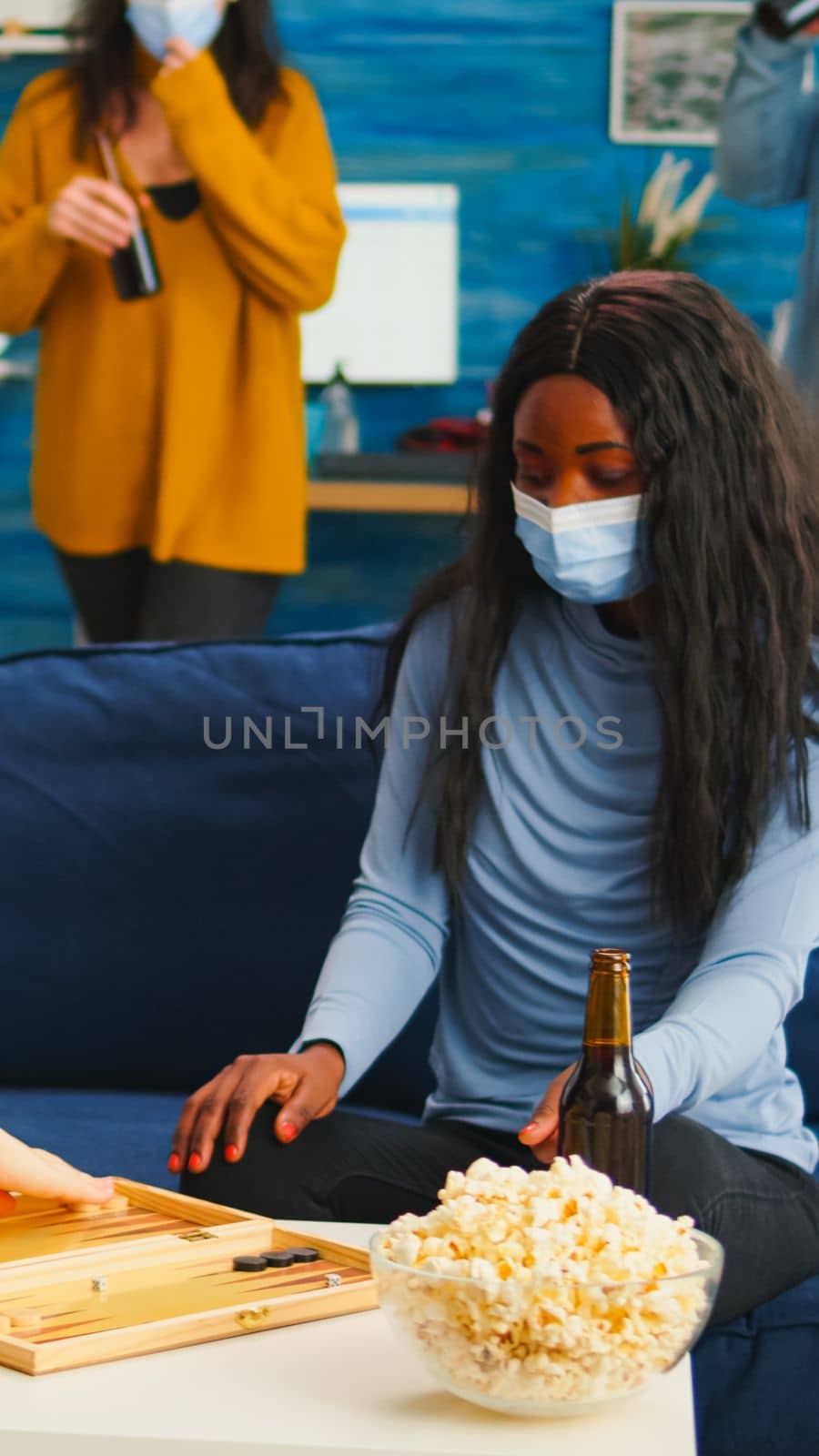 Multiethnical women playing backgammon wearing face mask as prevention for covid 19 spread during global pandemic sitting on sofa drinking beer and eating popcorn. Enjoying board games in outbreak