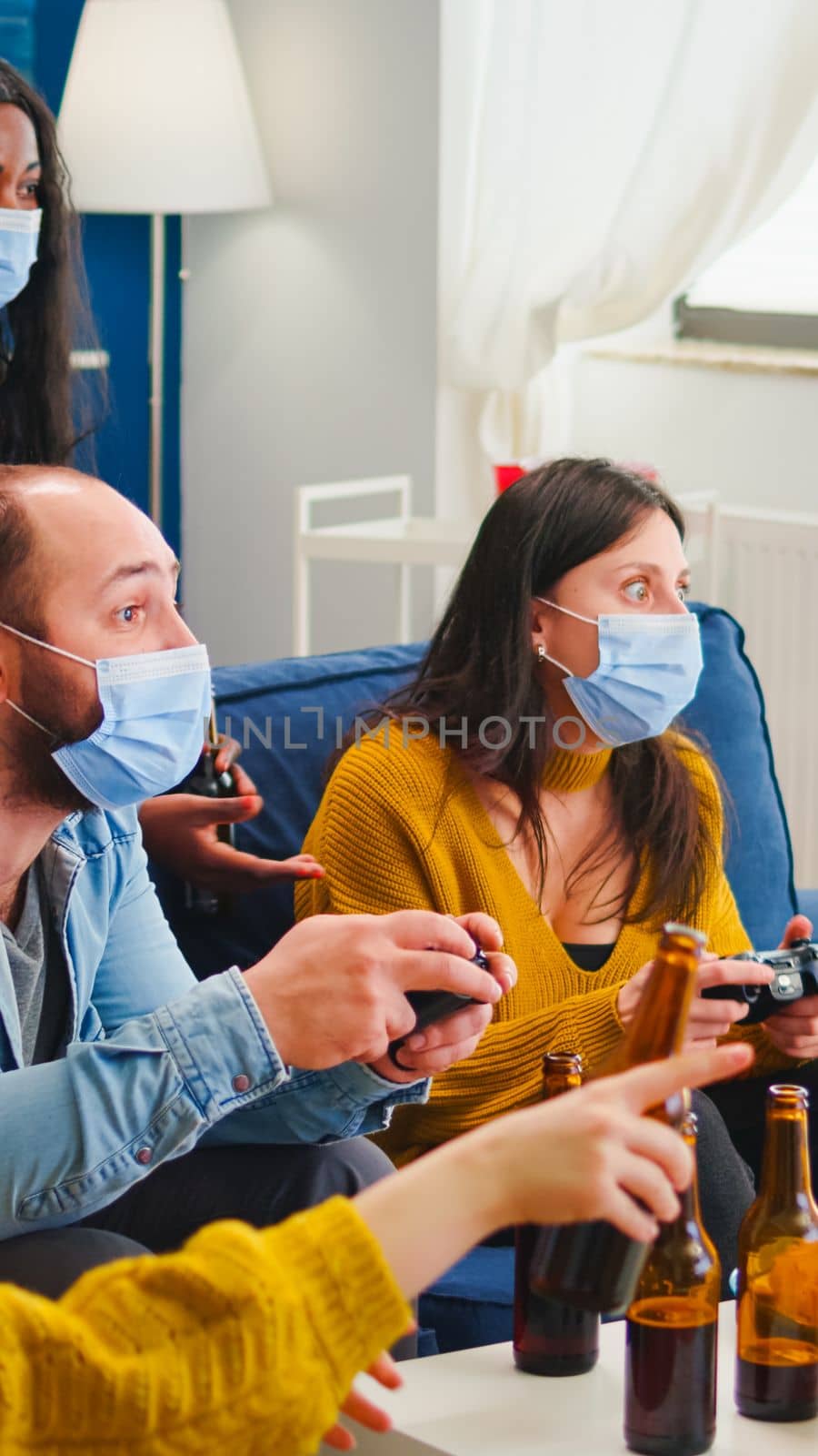 Sad friends because loosing playing video games with wireless controller wearing face mask to prevent getting sick with corona in time of social pandemic. Group of people enjoying time together.