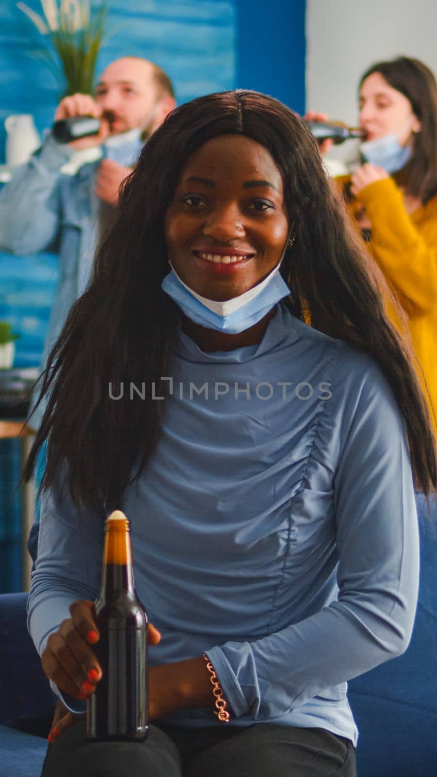 African woman keeping social distancing wearing face mask while meeting with friends to prevent spread of coronavirus holding beer bottle looking at camera sitting on couch, covid 19 outbreak