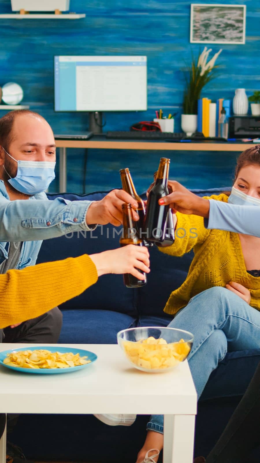 Mixed race friends clinking beer bottles hanging out keeping social distance having fun in living room during global pandemic. Diverse group of people celebrating with toast in outbreak enjoying time