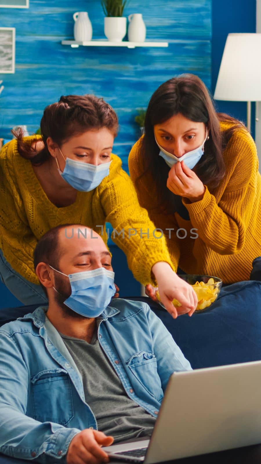 Cheerful happy multi ethnic friends looking at photo on laptop having fun together keeping social distance wearing face mask preventing coronavirus spreading. Diverse people enjoying new normal party