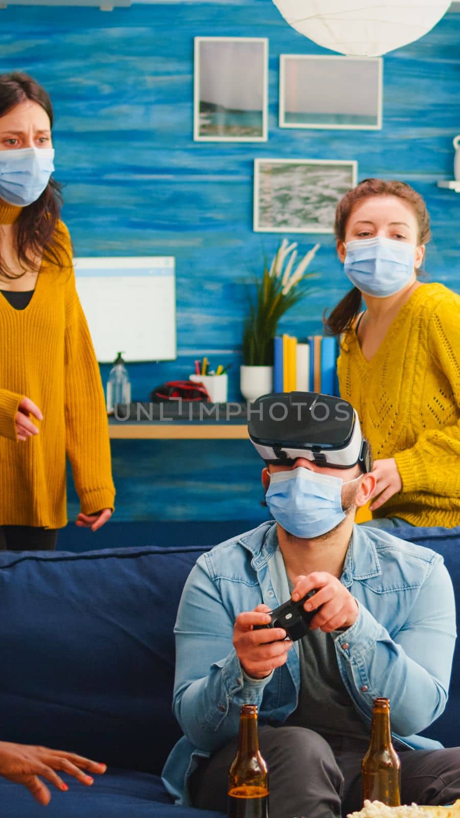 Group of multi ethnic friends having fun playing video games using virtual reality headset and joystick wearing face mask keeping social distancing. Diverse people enjoying at new normal party