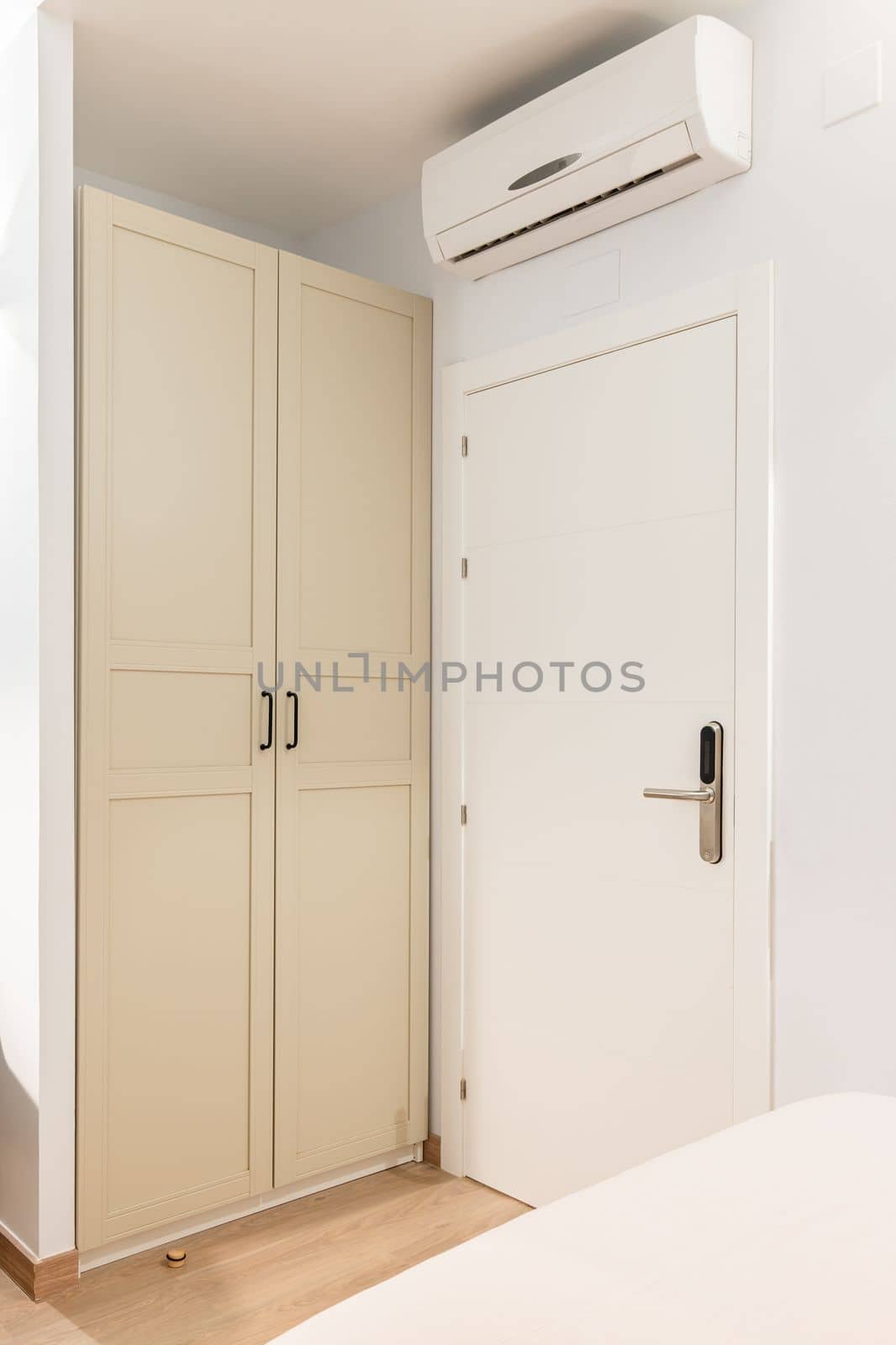 Closeup of a white wooden front door to a room next to a wardrobe. Wardrobe doors are painted beige and closed from prying eyes. Air conditioning above the door for pleasant coolness on hot nights. by apavlin