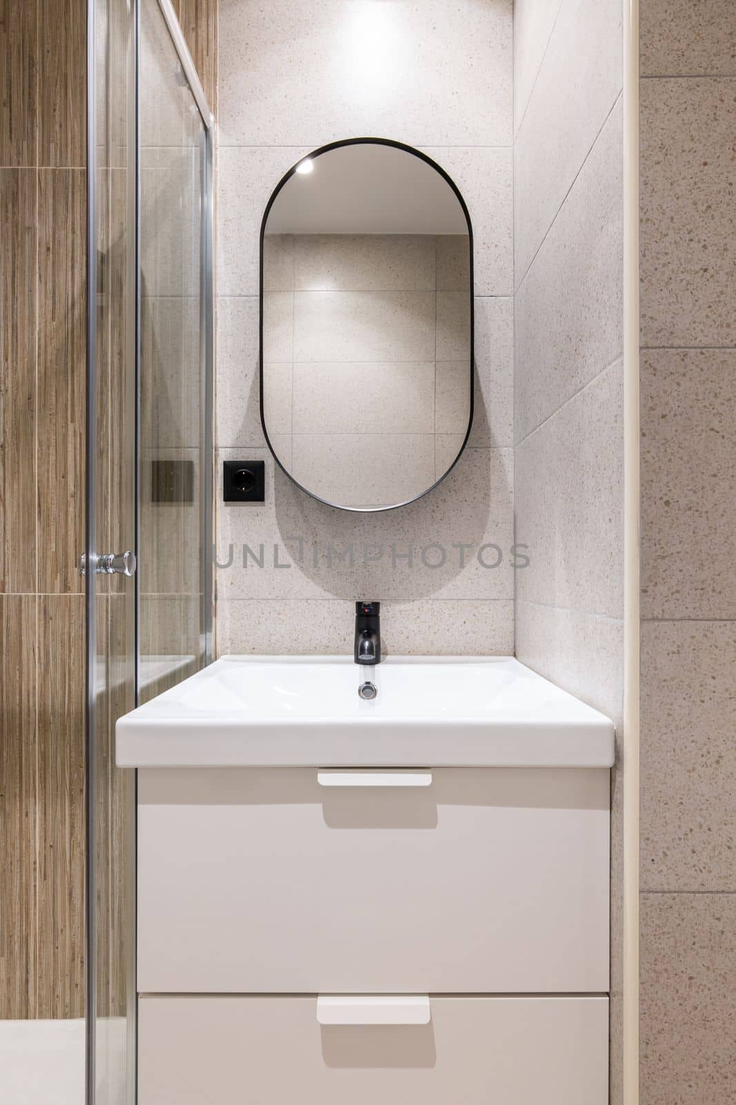 Closeup of a bathroom alcove with bright artificial lighting, with a white solid ceramic sink on a two-drawer vanity. Large modern oval mirror on the wall above the sink. by apavlin
