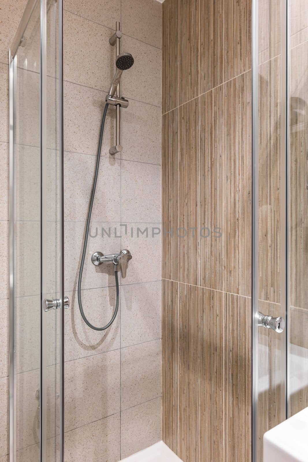 Shower area with marble tiles on the walls. On the wall is a faucet with a long hose and a shower head. Area is fenced with glass doors to protect against splashing water