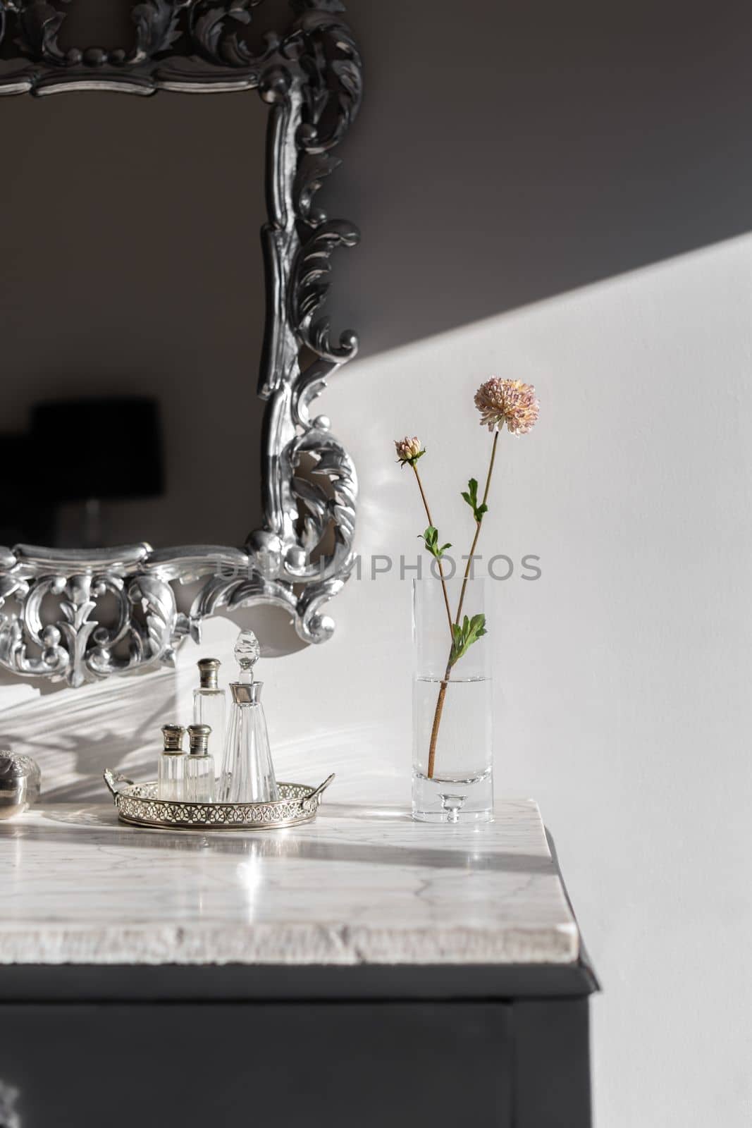 Part of dressing table with vase and flower bright by sunlight on white marble top. Mirror in silver frame with beautiful figured monograms. On table are empty bottles of fragrant perfume