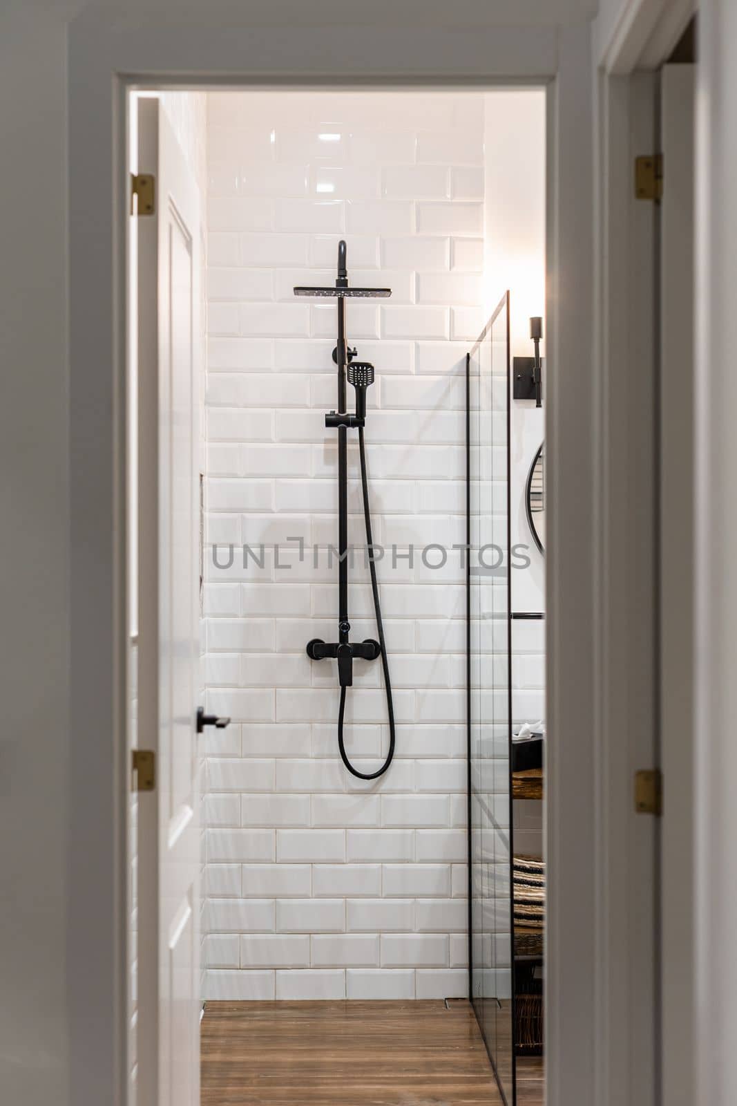 Looking at shower area in bathroom through doorway in hallway. White brick walls with paint reflecting bright light from lamps from ceiling. On wall is black metal faucet and glass railing on side. by apavlin