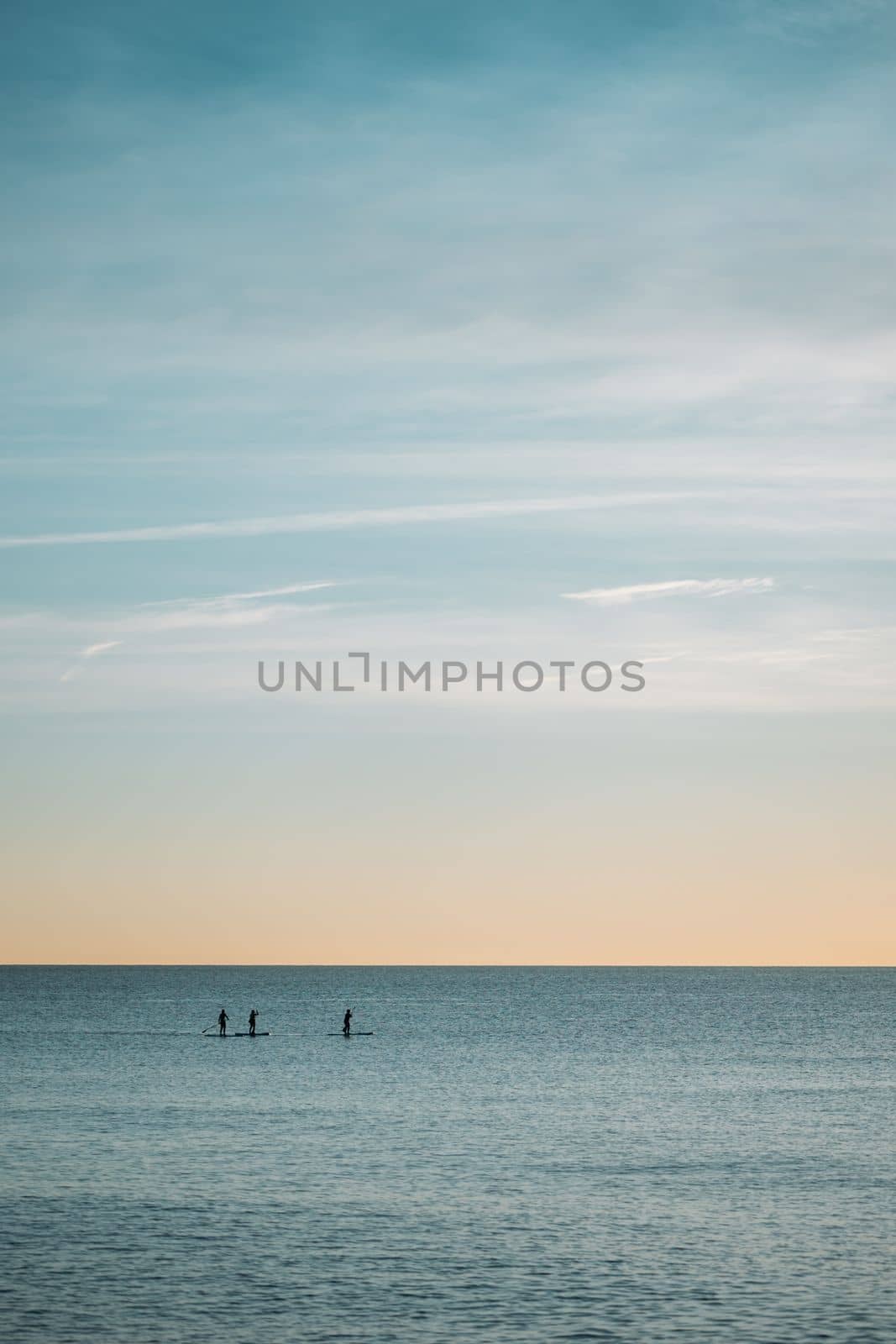 The horizon line where the clear blue cloudless sky and the calm ocean meet. In the distance, human silhouettes of paddle surfers with rowing oars in their hands. by apavlin