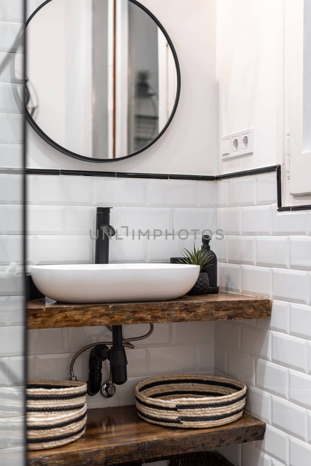 Bathroom with white brick walls, an oval washbasin on a wood-style marble top, a round mirror on a dark-framed wall reflects a doorway. Black ceramic faucet and wicker baskets under the sink. by apavlin