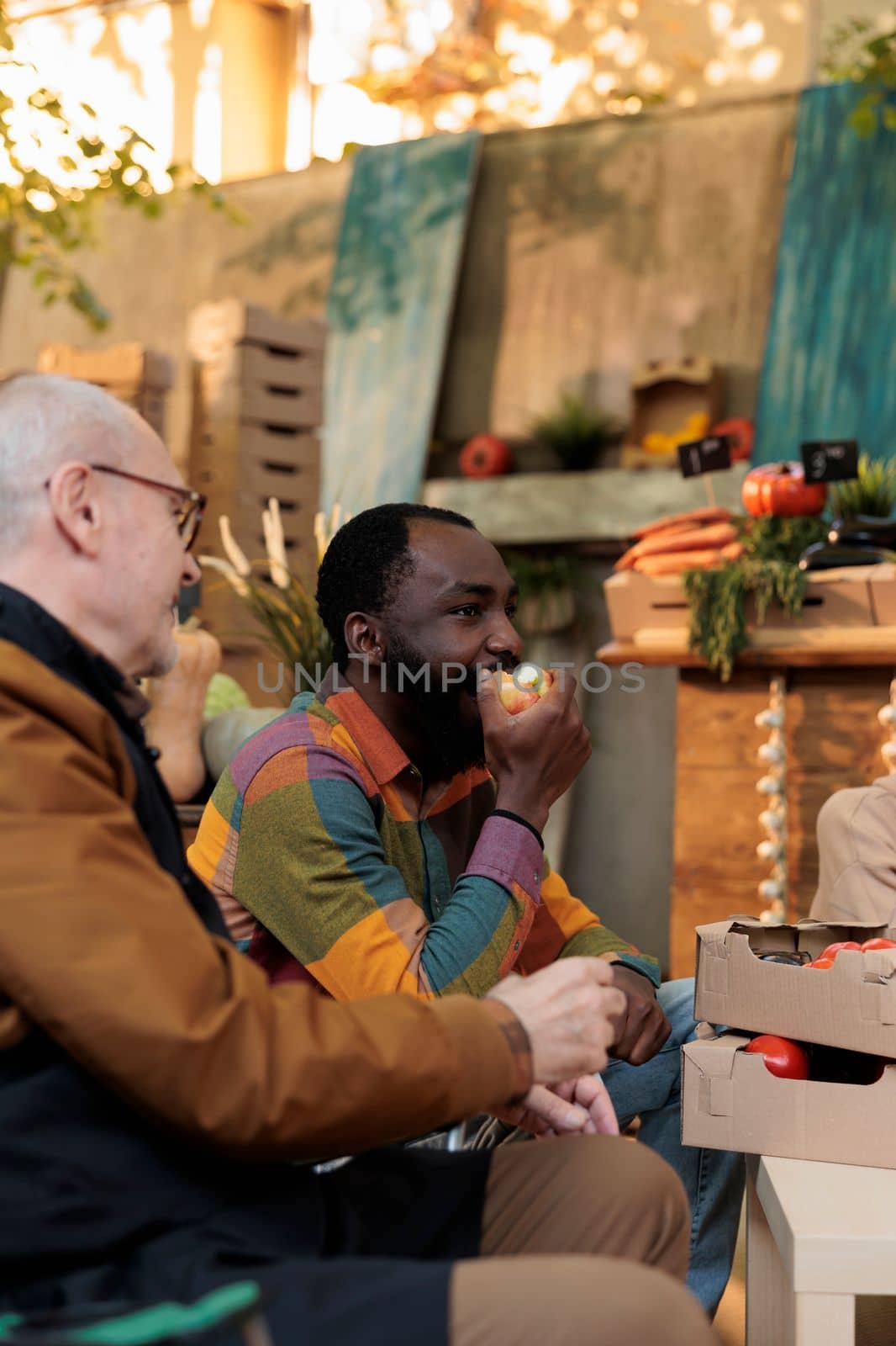 Young African American guy eating apple while sitting at table with friendly greengrocer near stand with organic produce, black man tasting fresh fruits and veggies from local farmers at autumn fair