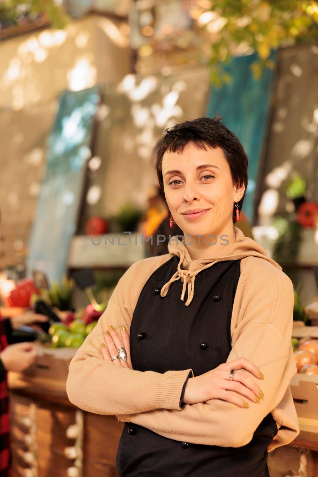 Woman stand owner wearing apron running farmers market stall, selling fresh organic produce directly to clients. Female greengrocer standing with crossed arms and looking at camera.