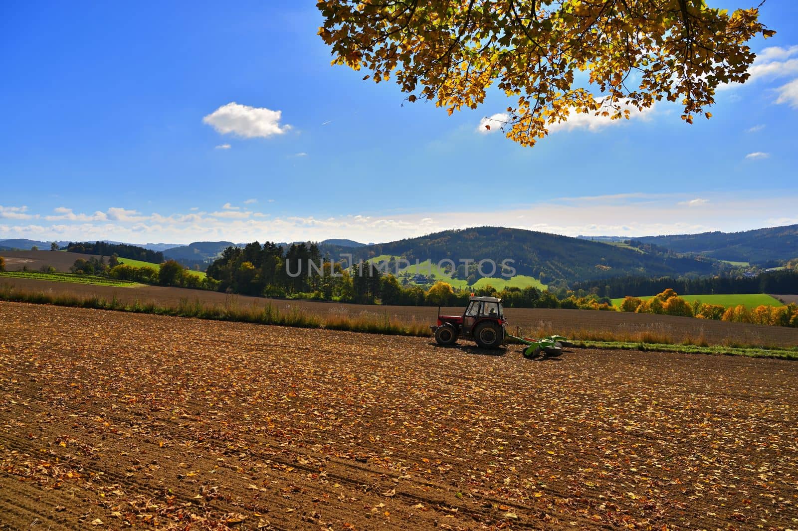 Tractor on the field. Beautiful autumn nature with landscape in the Czech Republic. Colorful trees with blue sky and sun. Background for autumn and agriculture.