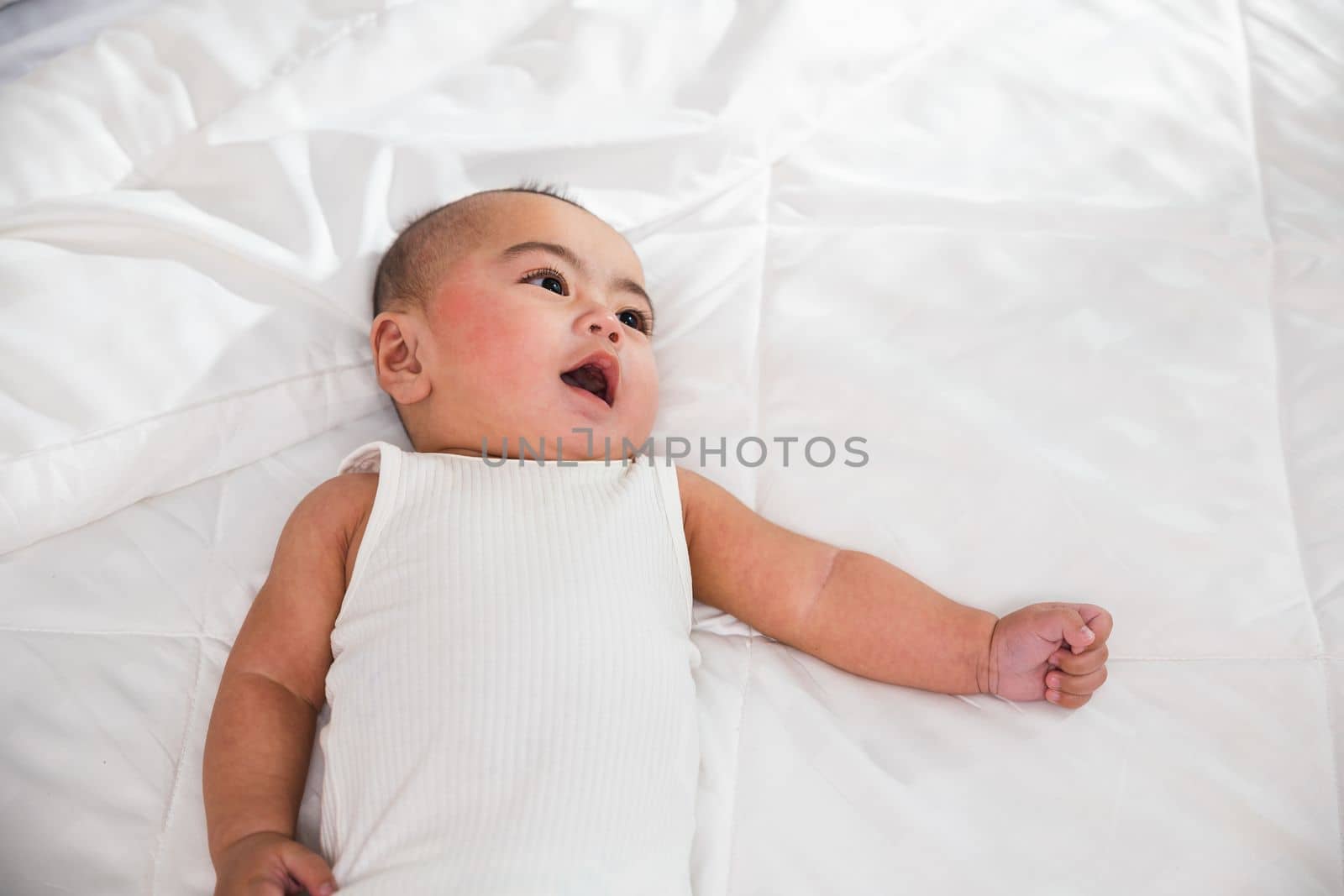 Asian cute little baby on bed under soft blanket, baby wearing bodysuit lying on white beedsheets in bedroom, smiling infant child at home