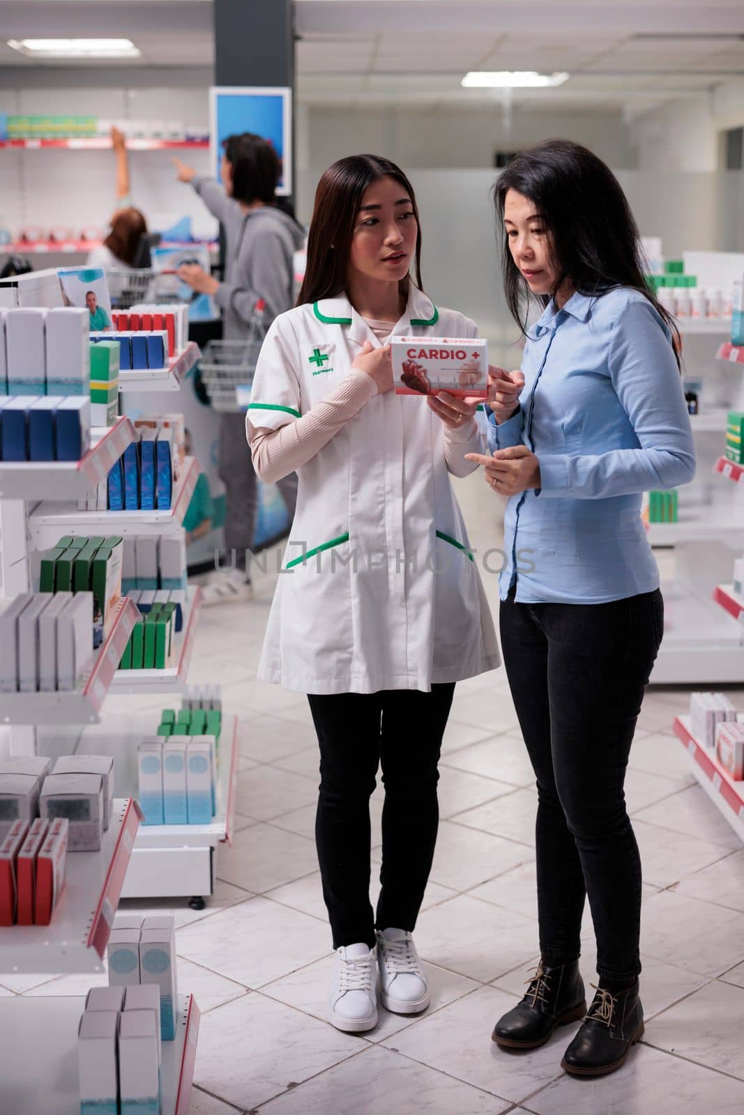 Asian assistant and client examining cardiology pills box from drugstore shelves, buying medicinal supplements in pharmacy. Customer talking about pharmaceutical products supplies.
