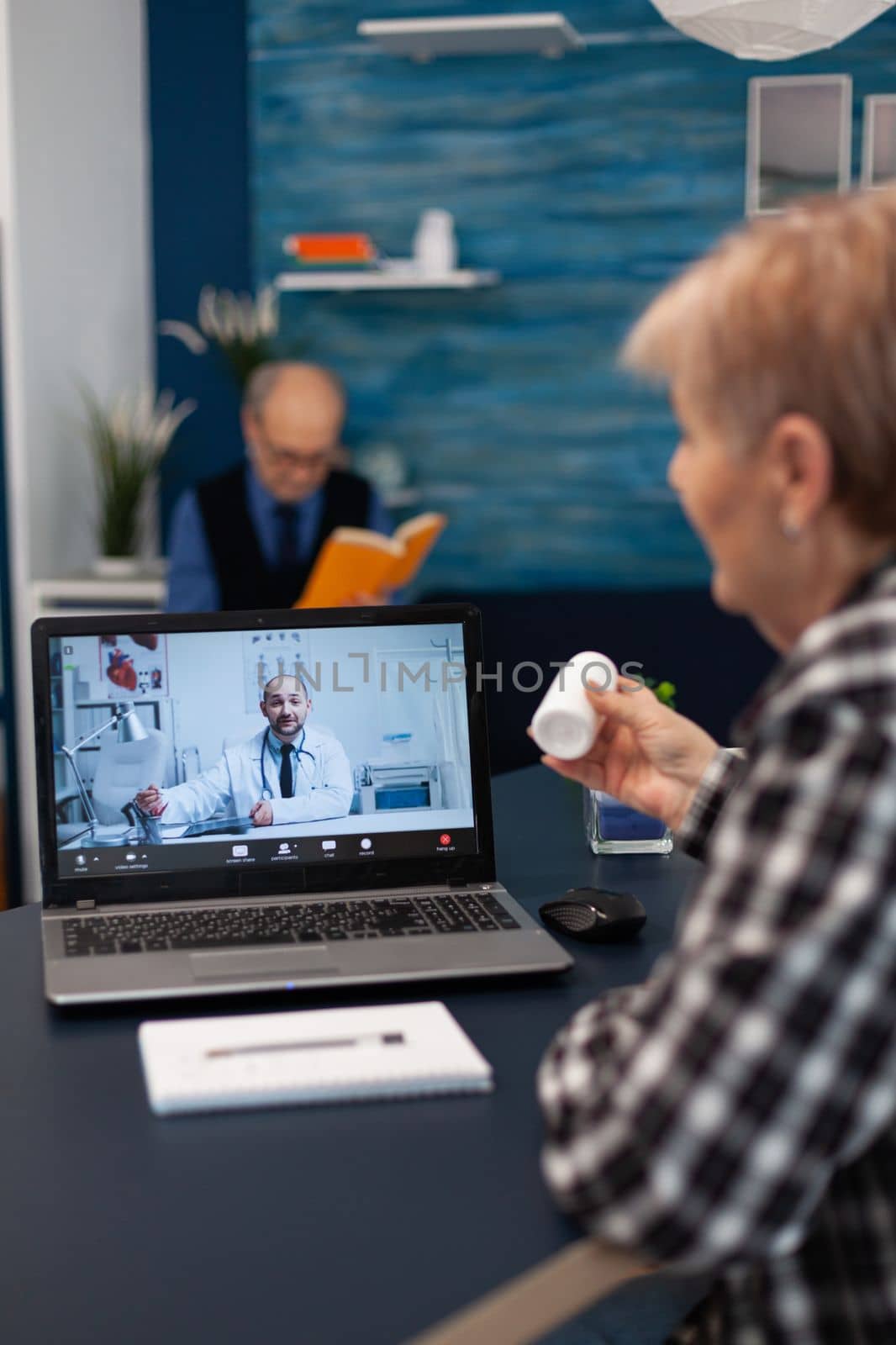 Senior pensioner listening doctor talking about treatment during online consultation. Elderly woman discussing with healthcare practitioner in the course of remote call and husband is reading a book on sofa.