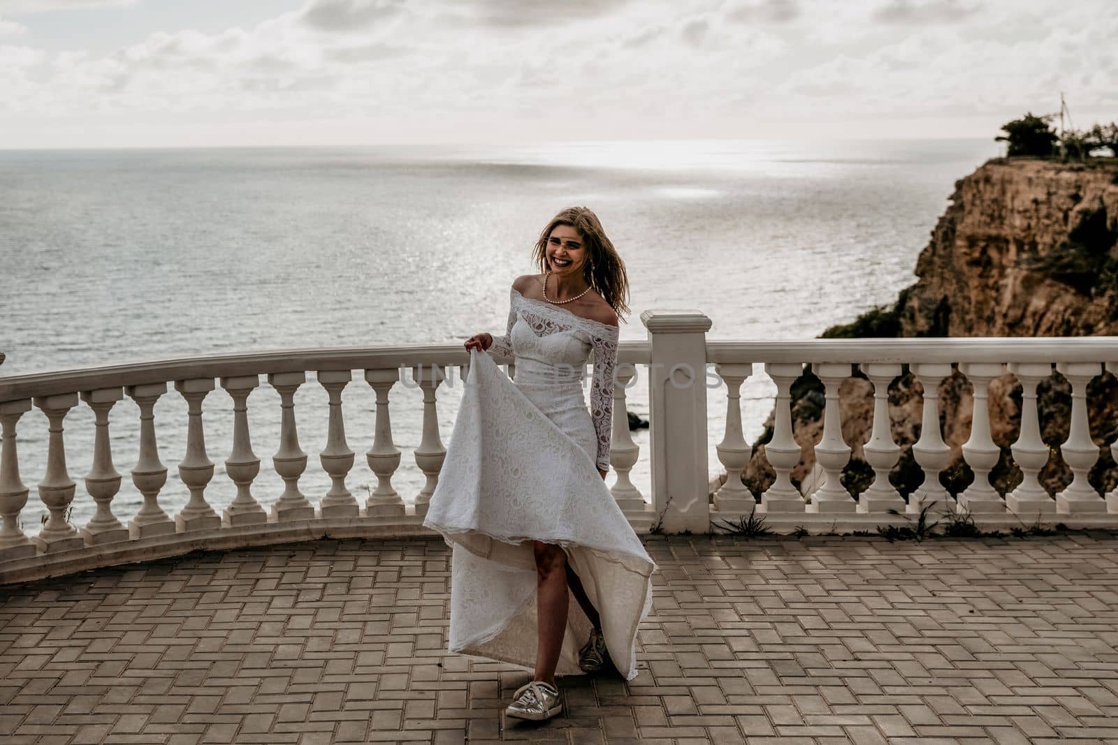 Romantic beautiful bride in white dress posing with sea and mountains in background. Stylish bride standing back on beautiful landscape of sea and mountains on sunset