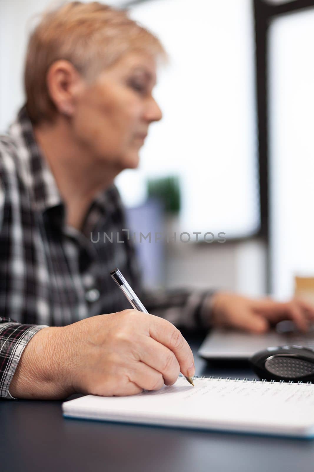 Close up of senior business woman writing notes on notebook. Elderly woman in home living room using moder technoloy laptop for communication sitting at desk indoors.