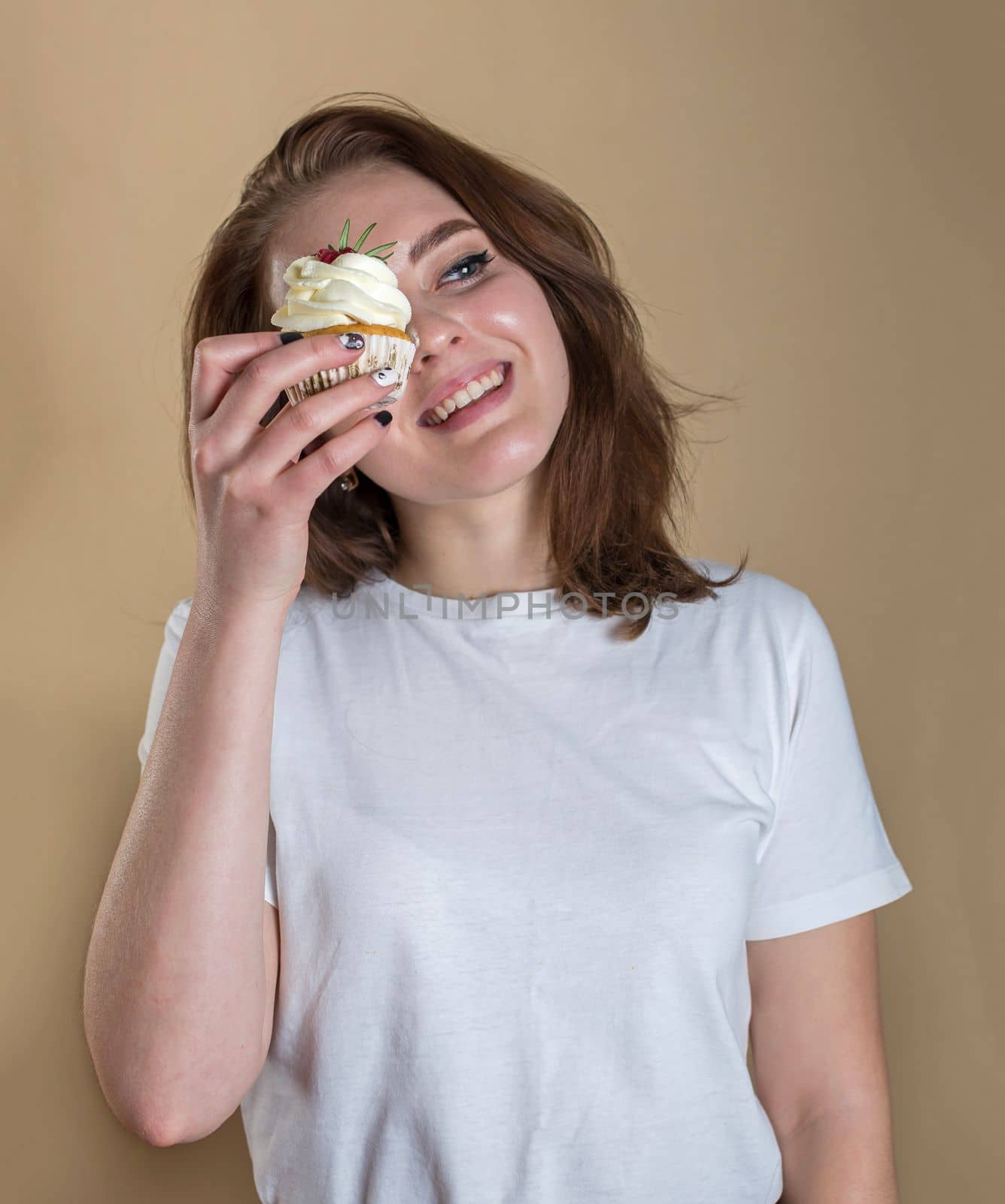 Portrait of a happy young girl with bright makeup on a beige background, eating cupcake