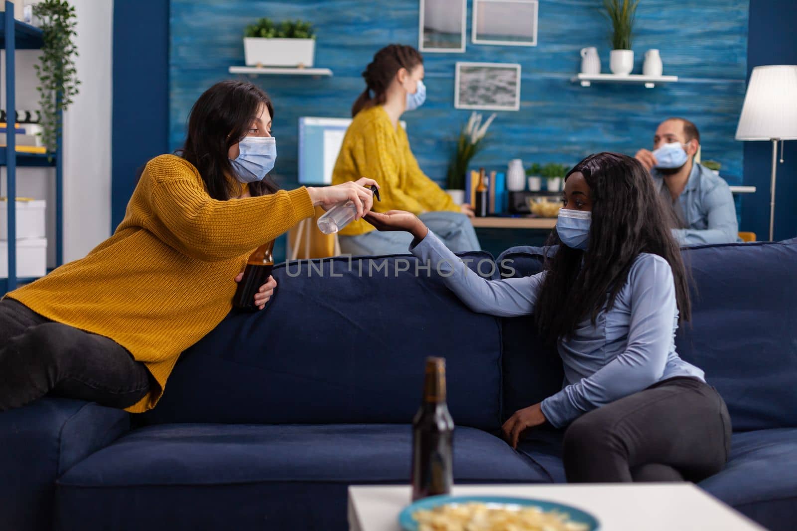 Group of multi ethnic people using hand sanitizer gel to prevent covid19 spread spending time together in living room sitting on sofa wearing face mask. Diverse people socializing keeping social distance