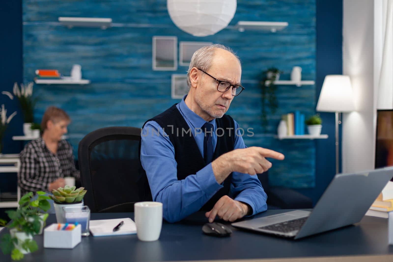 Thoughtfull senior businessman pointing at laptop whille working from home. Elderly man entrepreneur in home workplace using portable computer sitting at desk while wife is holding tv remote.