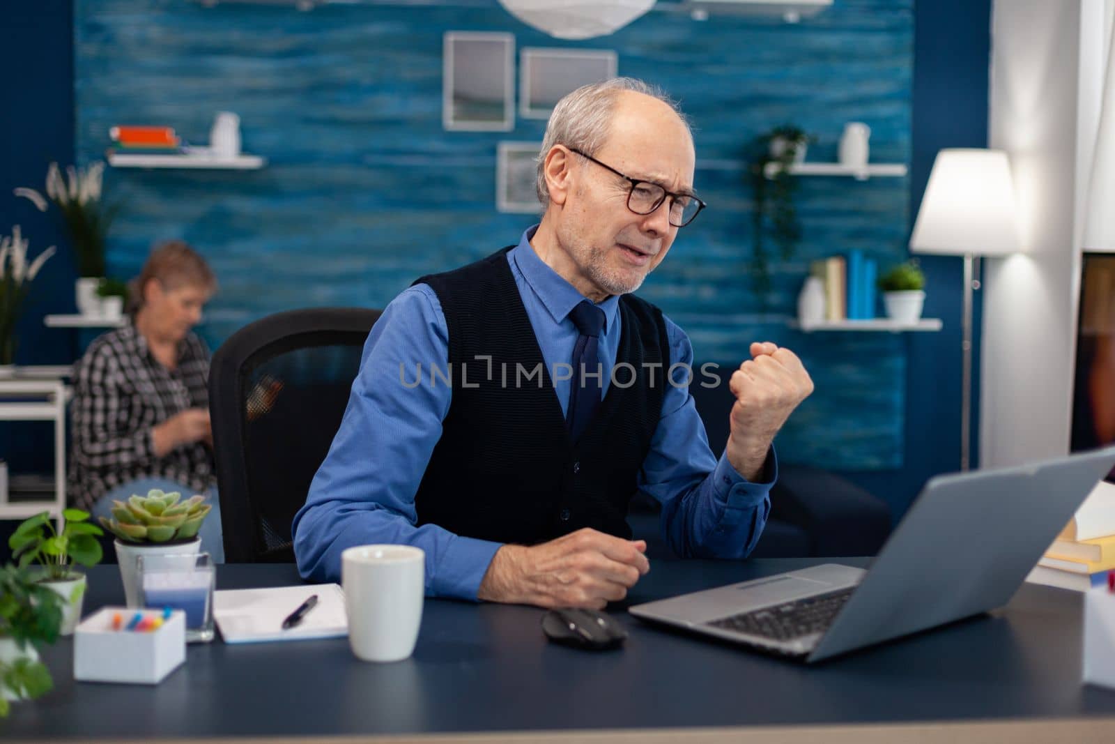 Excited senior man celebrating good news while working on laptop from home office. Elderly man entrepreneur in home workplace using portable computer sitting at desk while wife is reading a book sitting on sofa.