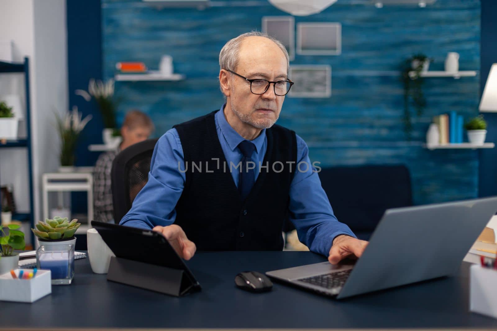 Senior manager working on presentation using laptop and tablet pc sitting at office. Elderly man entrepreneur in home workplace using portable computer sitting at desk while wife is reading a book sitting on sofa.