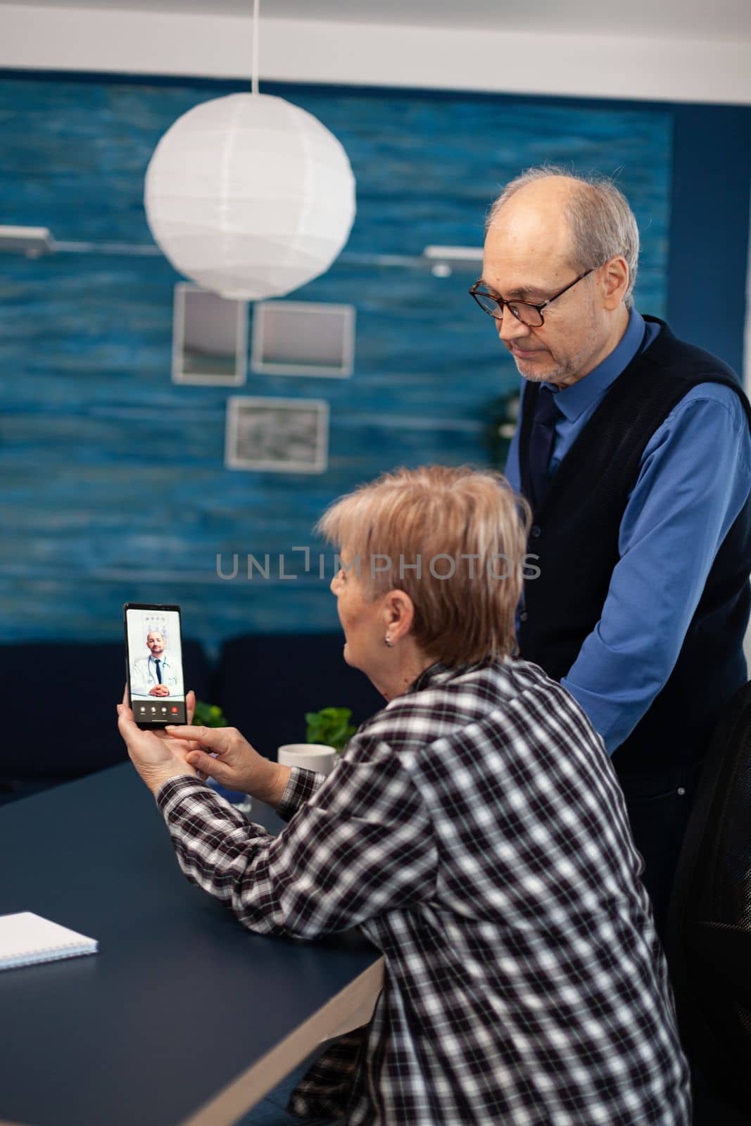 Senior sick man and woman talking with doctor during video call. Elderly woman and woman discussing with healthcare practitioner in the course of remote call and husband is reading a book on sofa.