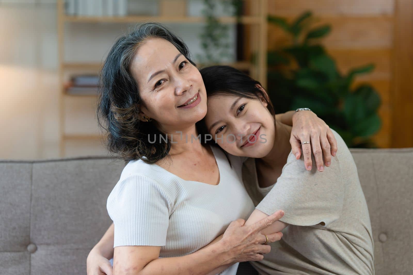 Happy senior Asian woman and her adult daughter are cuddling on the couch.