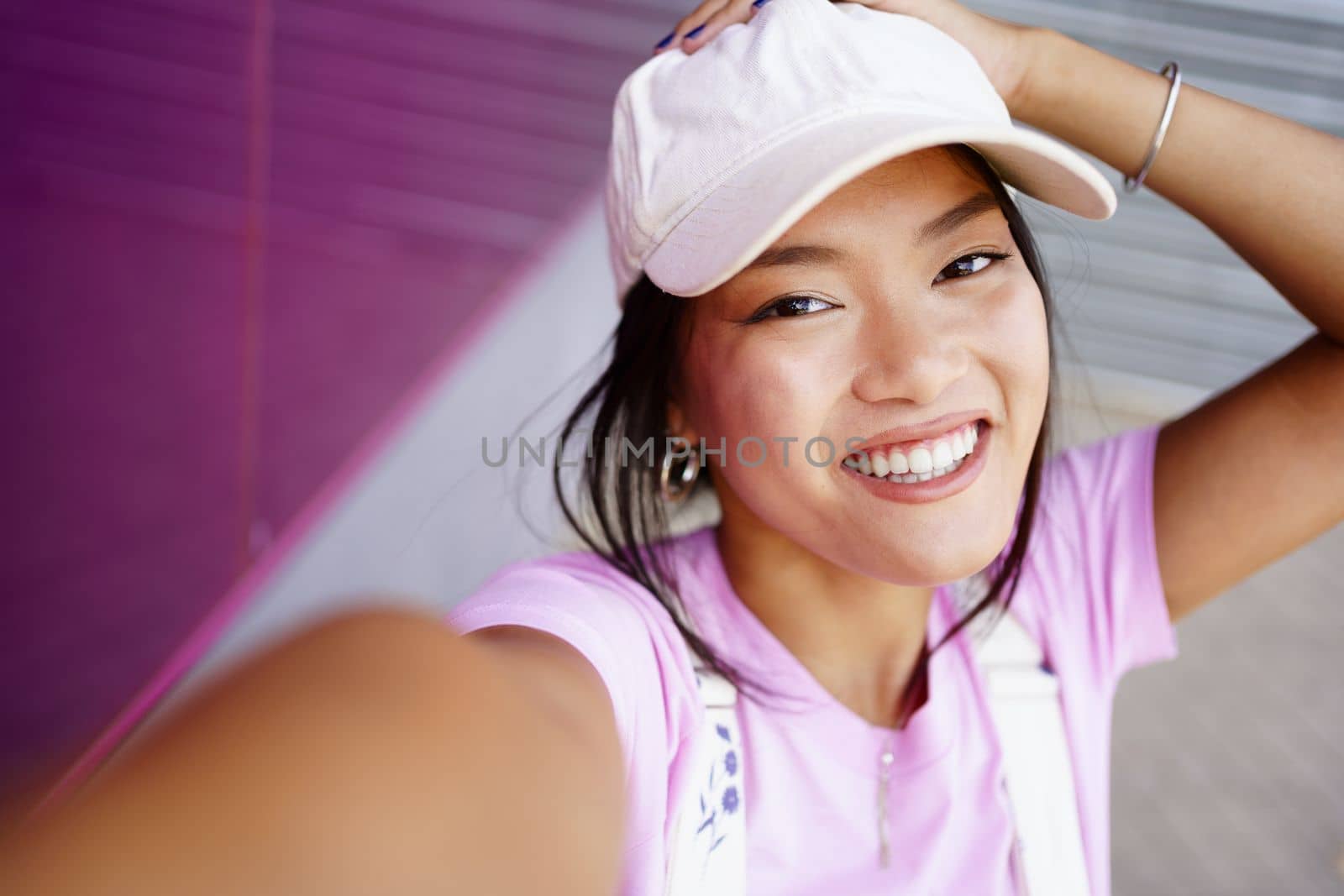 Positive young Asian woman in stylish clothes touching head and looking at camera with smile while taking selfie on city street in daytime
