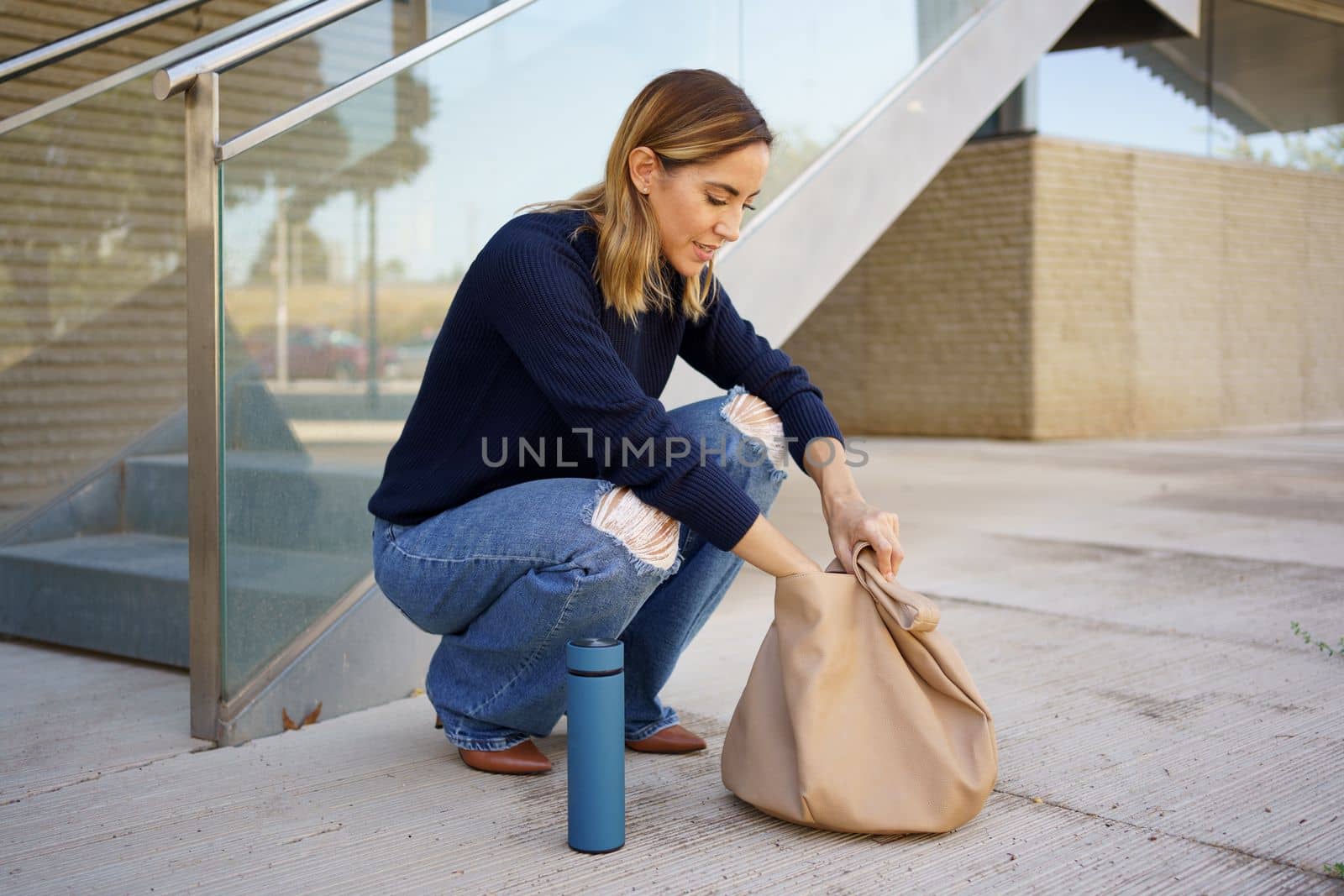 Full body female in stylish clothes sitting on haunches near steps and searching for keys inside bag outside modern apartment building