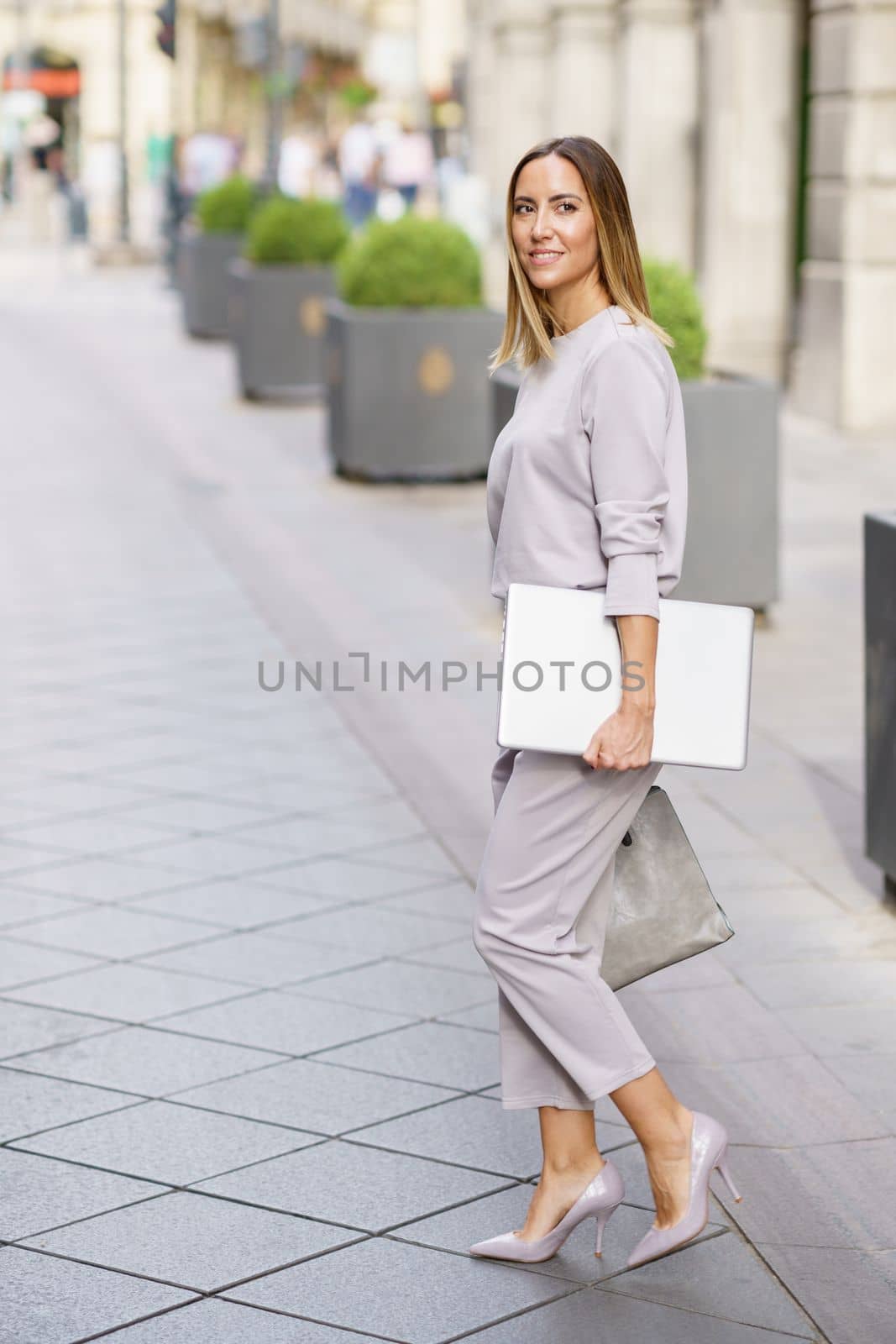 Side view full body of self assured young female entrepreneur with blond hair, in classy clothes and high heels with laptop in hand smiling while walking on paved street in city