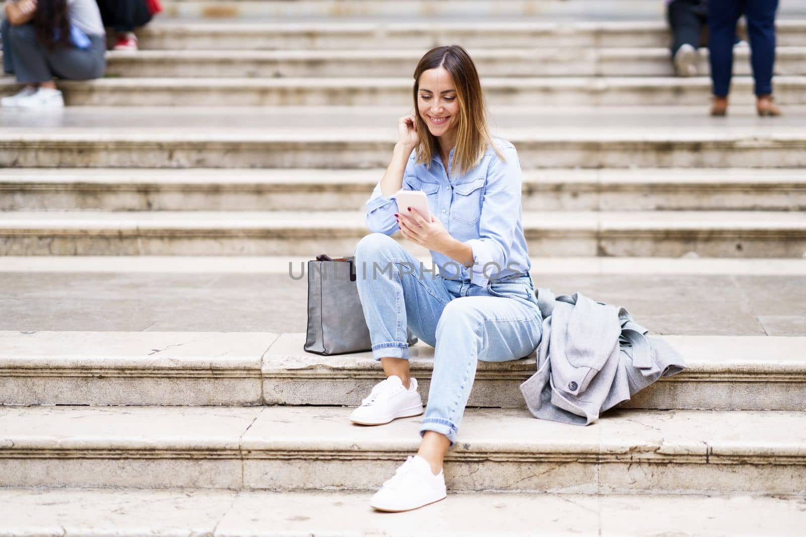 Full body glad adult woman in casual clothes touching hair and watching video on smartphone, while sitting on steps near stylish bag and coat in daytime on city street