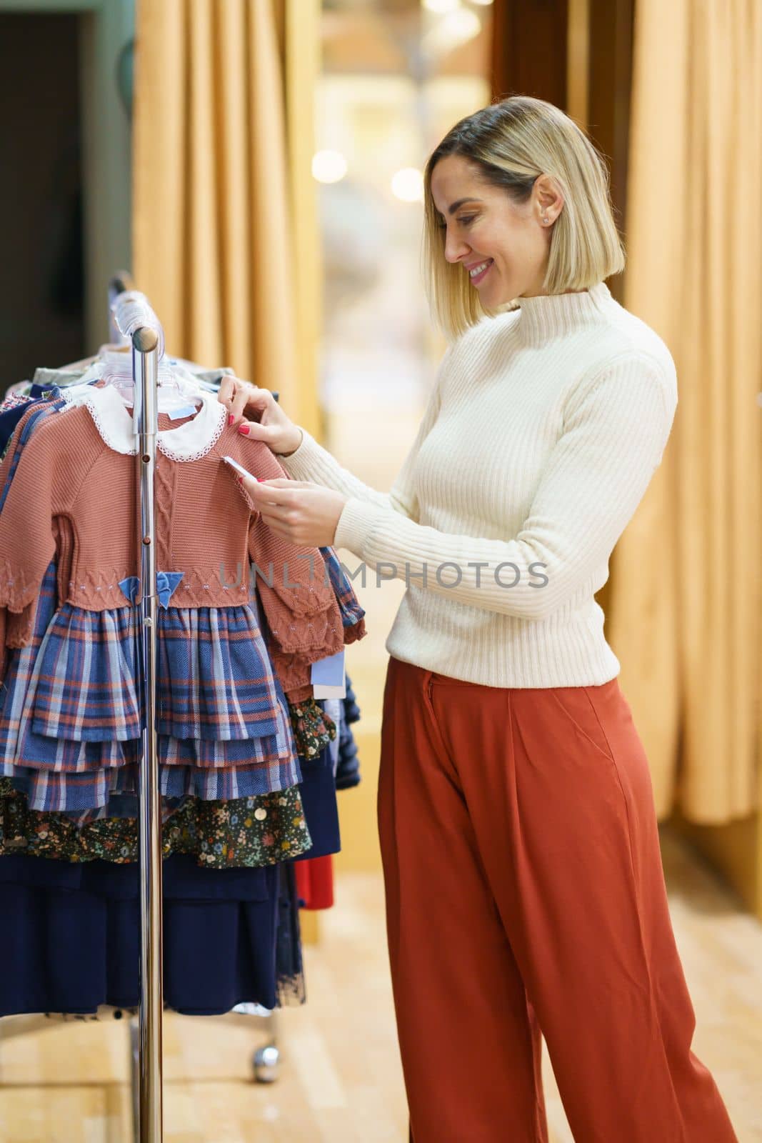 Smiling female customer looking at price of clothes hanging on rail in child store during shopping in mall