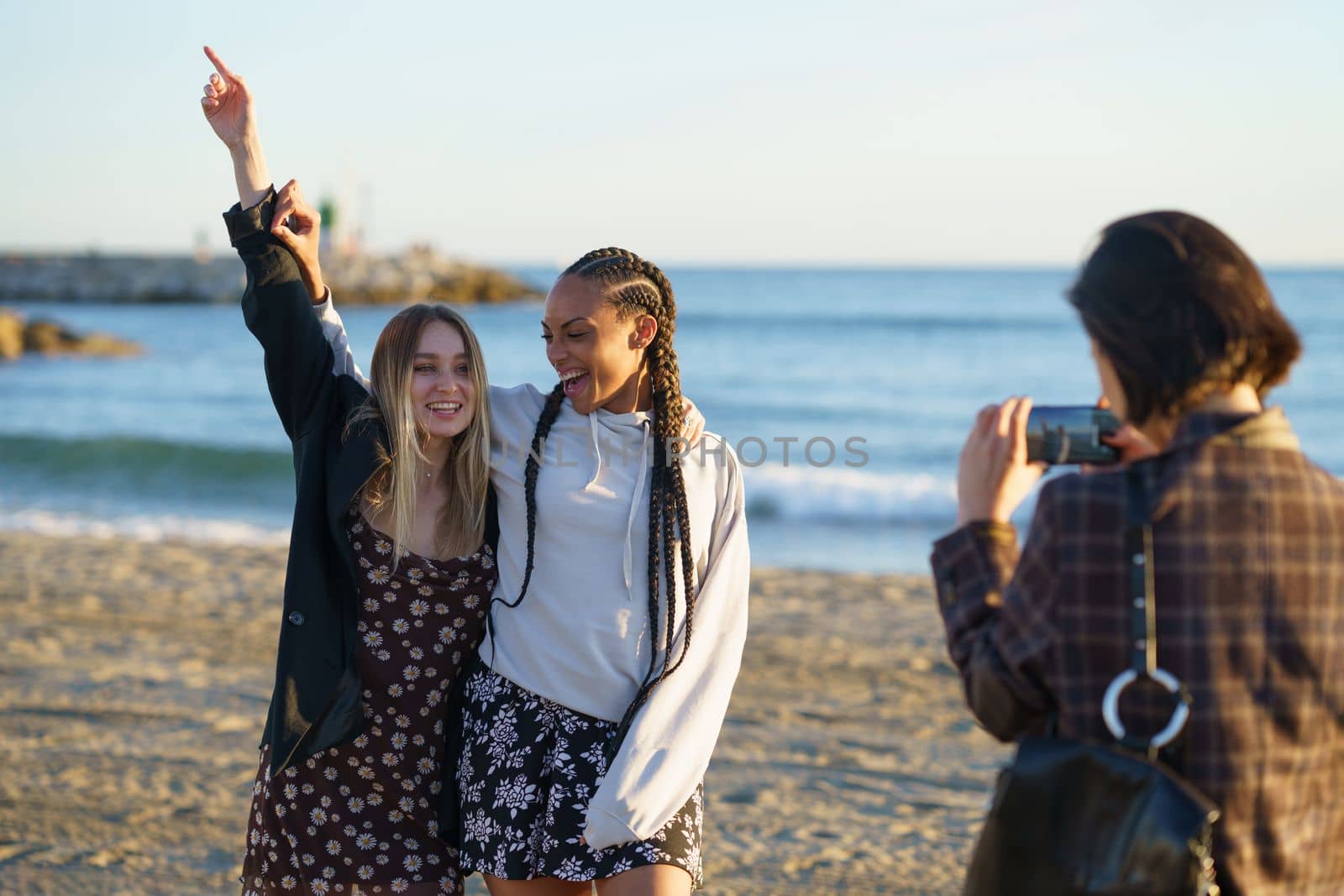 Unrecognizable woman with smartphone taking photo of smiling diverse female friends standing with raised arm on sandy coast near sea