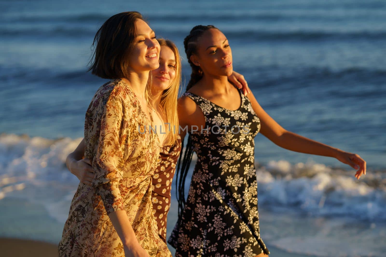 Positive multiracial female friends hugging and strolling together on seashore along waving sea on sunny summer day in coastal terrain