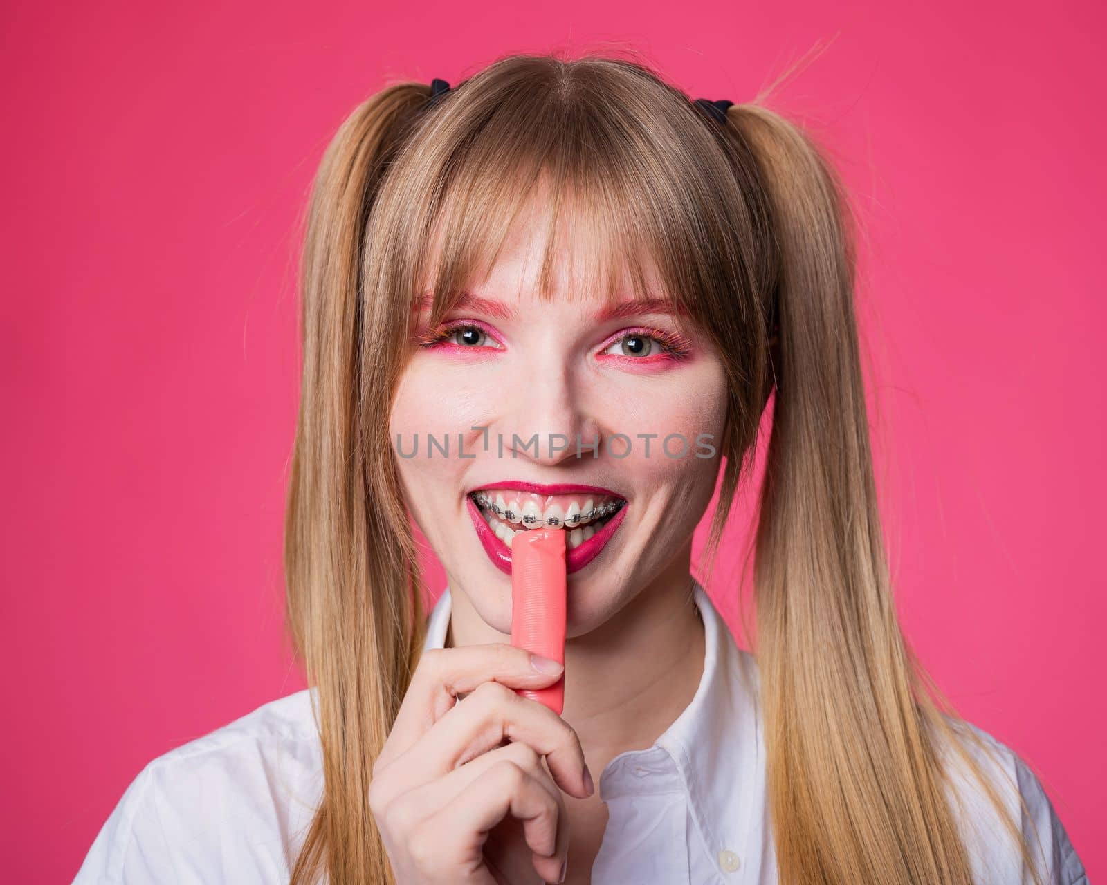 Portrait of a young woman with braces and bright makeup chewing gum on a pink background. by mrwed54